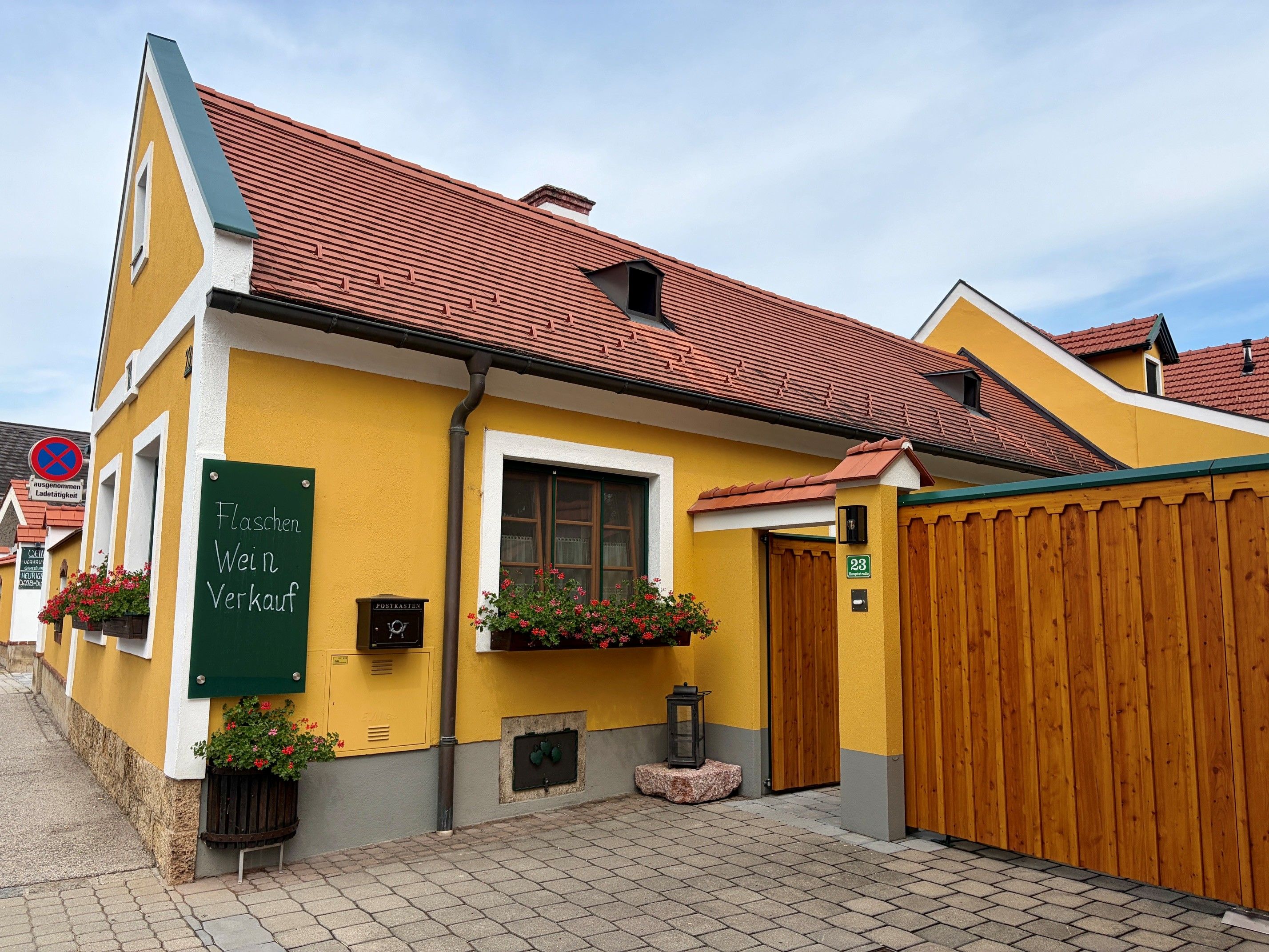Gelbes Gebäude mit rotem Dach und Schild 'Flaschen Wein Verkauf'. Blumenkästen an den Fenstern und Holztor im Vordergrund.
