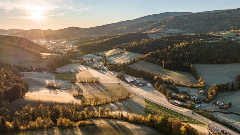 Luftaufnahme einer ländlichen Landschaft bei Sonnenaufgang mit Feldern, Wäldern und vereinzelten Gebäuden.