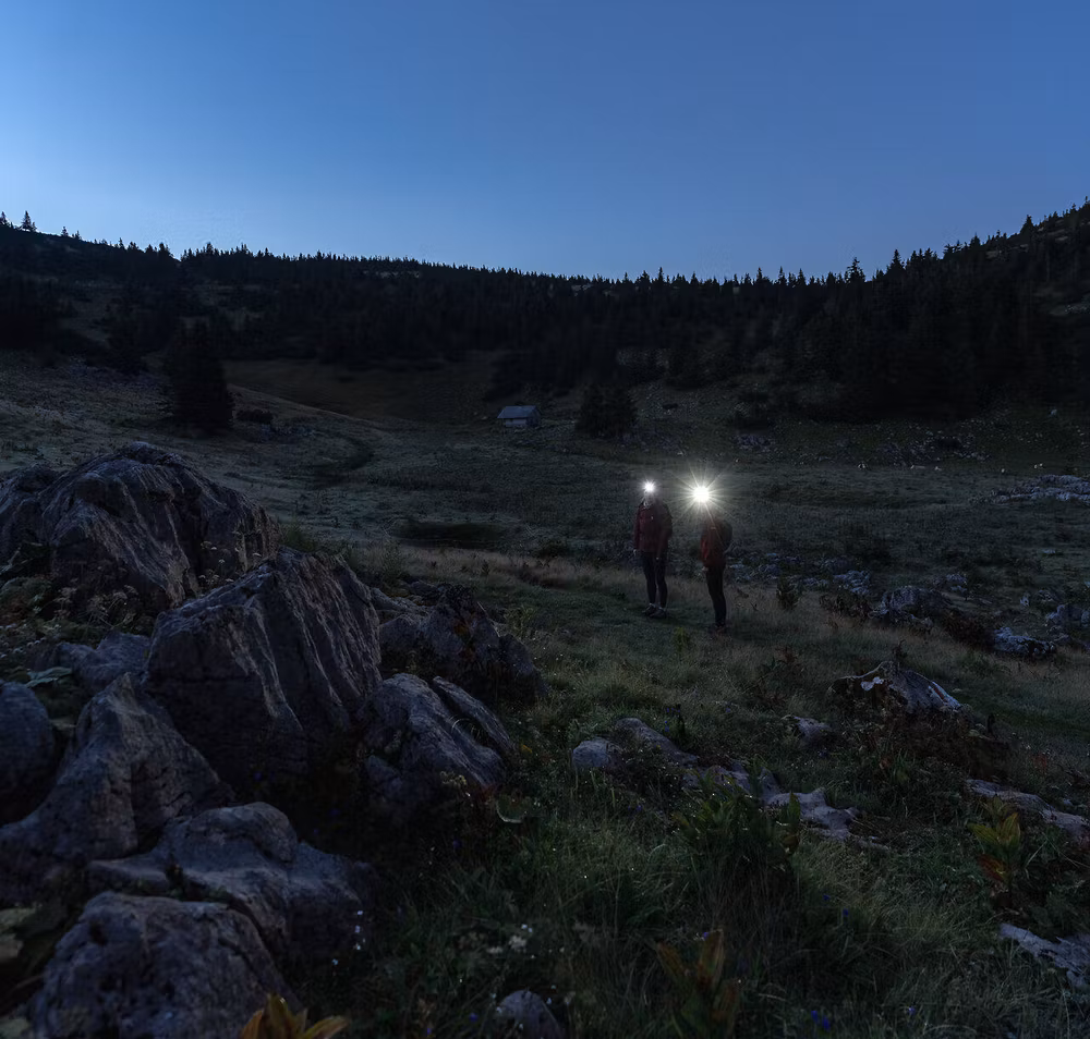 In der sanften Dämmerung erstrahlen die Ybbstaler Alpen in einem magischen Licht. Zwei Wanderer, ausgestattet mit Stirnlampen, erkunden die unberührte Natur und genießen die frische Morgenluft, während die ersten Sonnenstrahlen den Horizont küssen.
