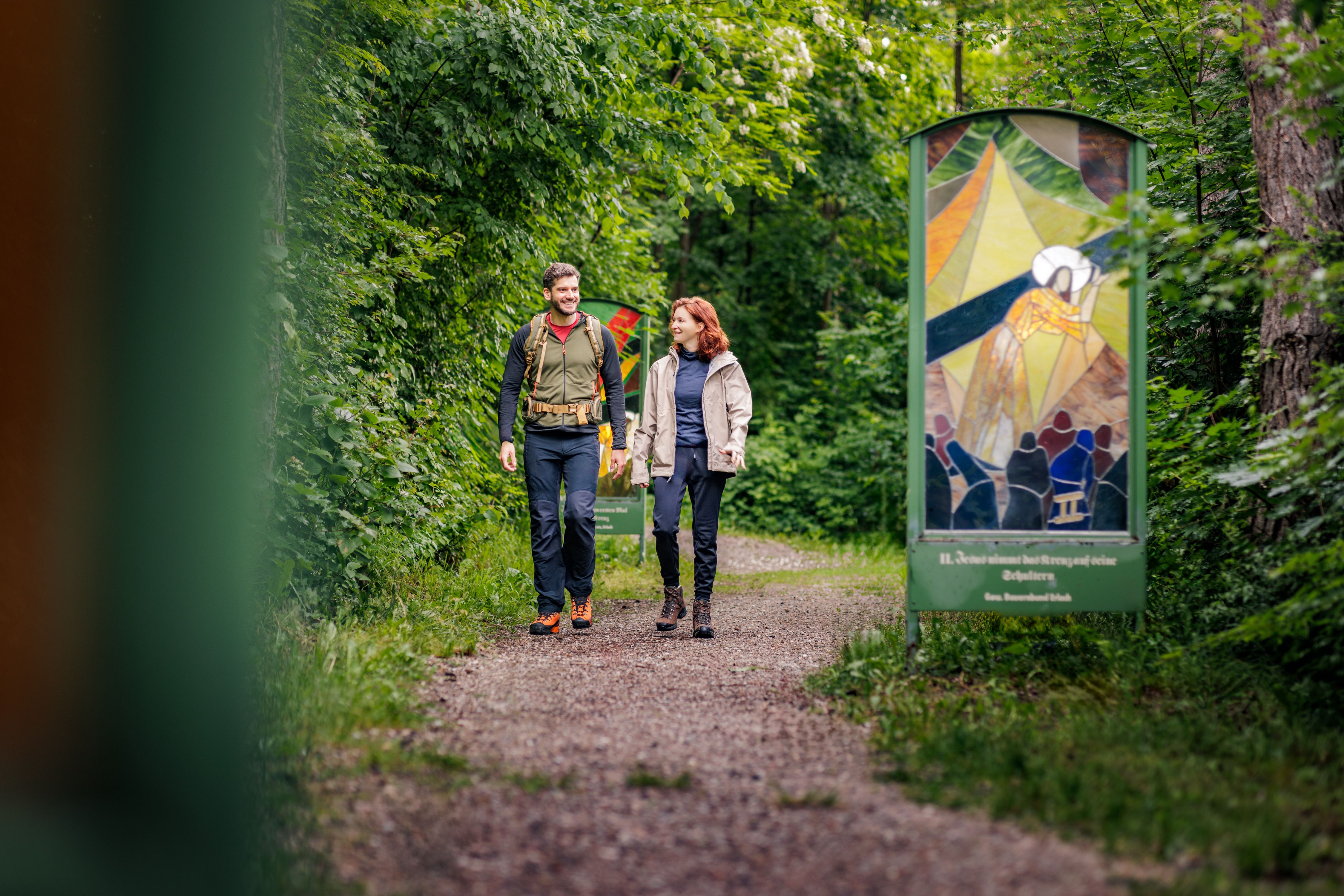 Zwei Personen wandern auf einem Waldweg mit gläsernen Kunstwerken am Rand.