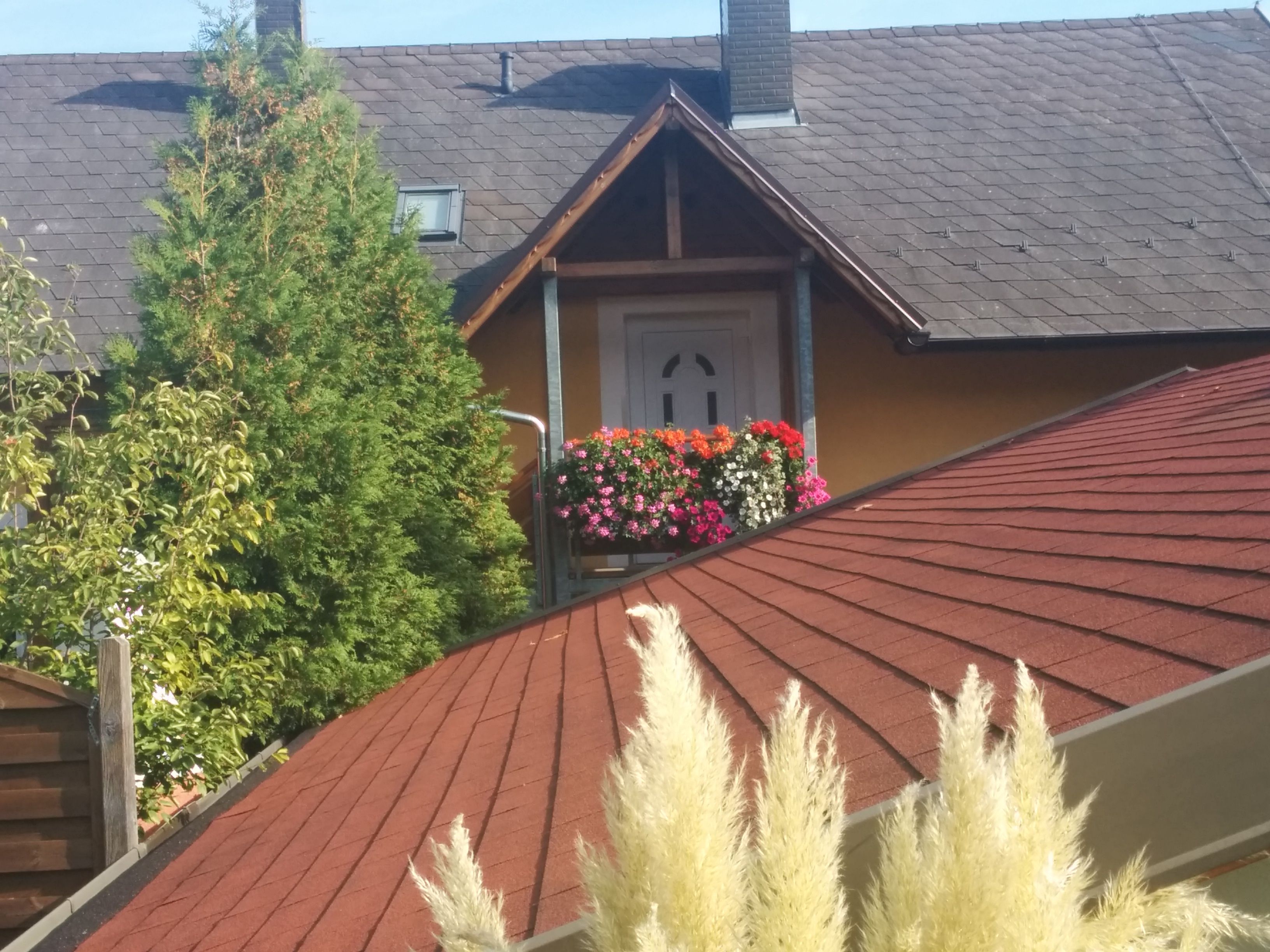 Hausdach mit rotem Schindeldach, Balkon mit Blumen, Baum im Hintergrund.