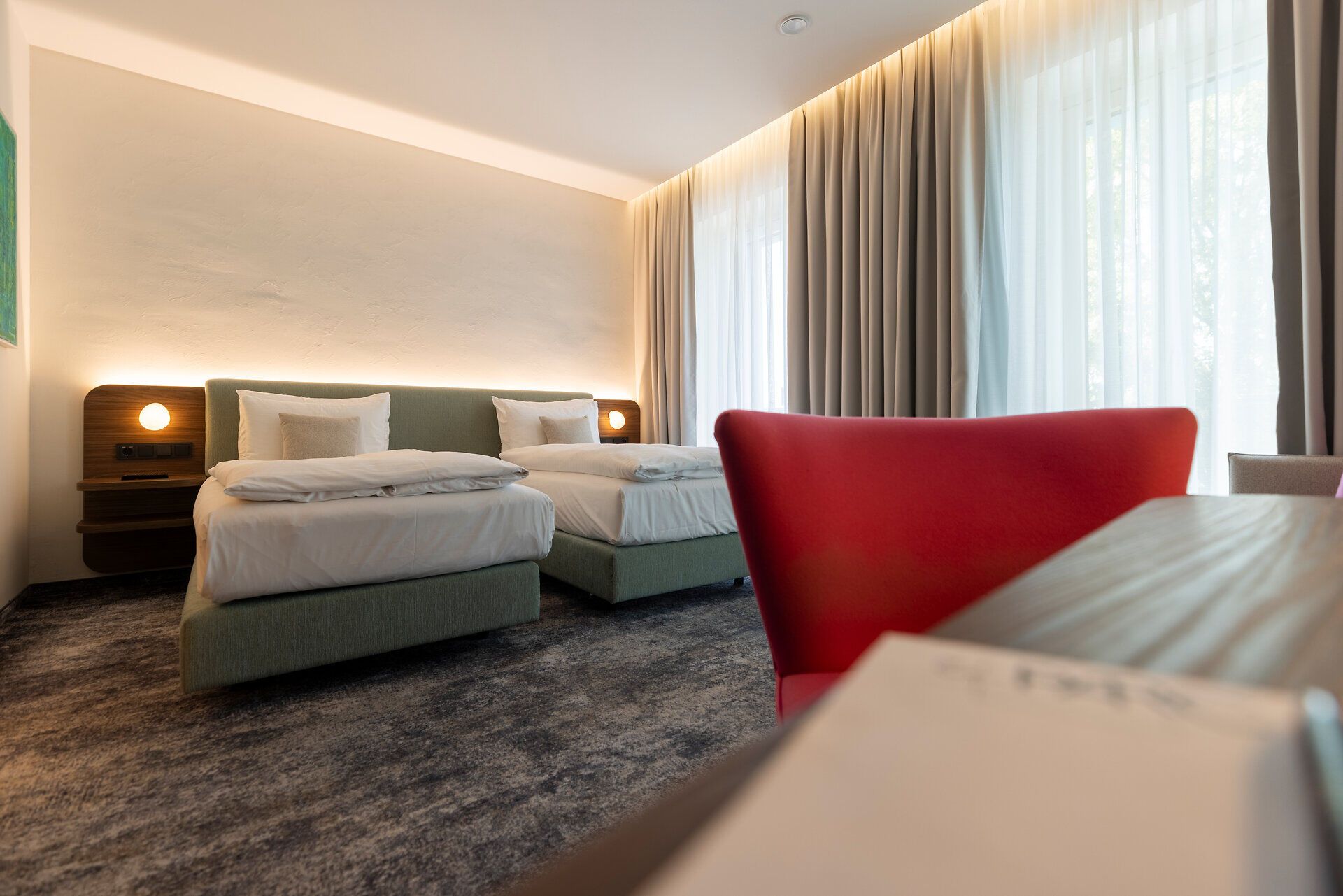 Im modern eingerichteten Zimmer mit großen Fenstern vom Hotel leParc befinden sich zwei Betten sowie ein Tisch mit rotem Sessel. 