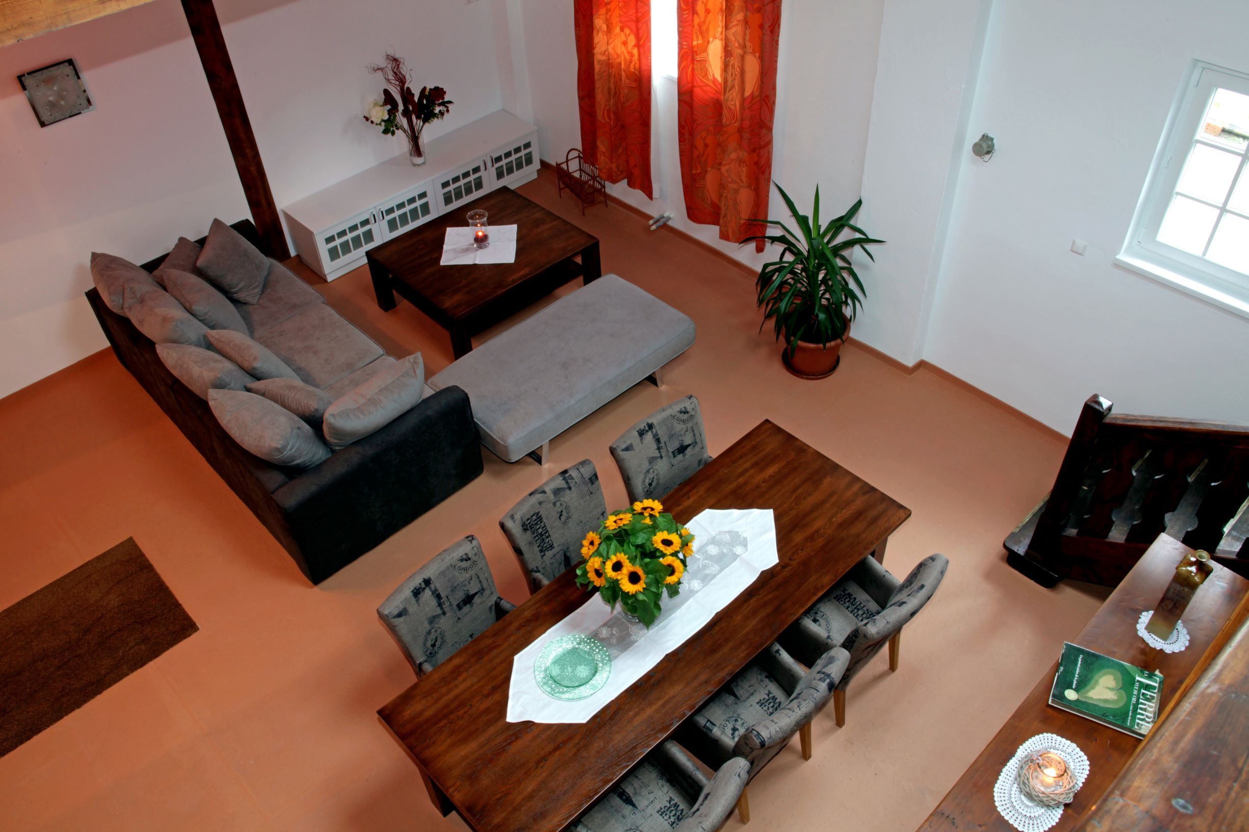 Blick von oben auf ein Wohnzimmer mit grauem Sofa, Esstisch mit Sonnenblumen, roten Vorhängen und einer Pflanze.