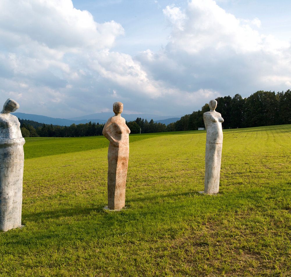 Drei stilisierte Figuren aus Stein stehen majestätisch auf einer weitläufigen Wiese, umgeben von sanften Hügeln und einem beeindruckenden Himmel. Diese Kunstwerke laden dazu ein, innezuhalten und die harmonische Verbindung zwischen Natur und Kunst zu genießen. Ein Ort der Ruhe und Inspiration, der die Seele berührt.