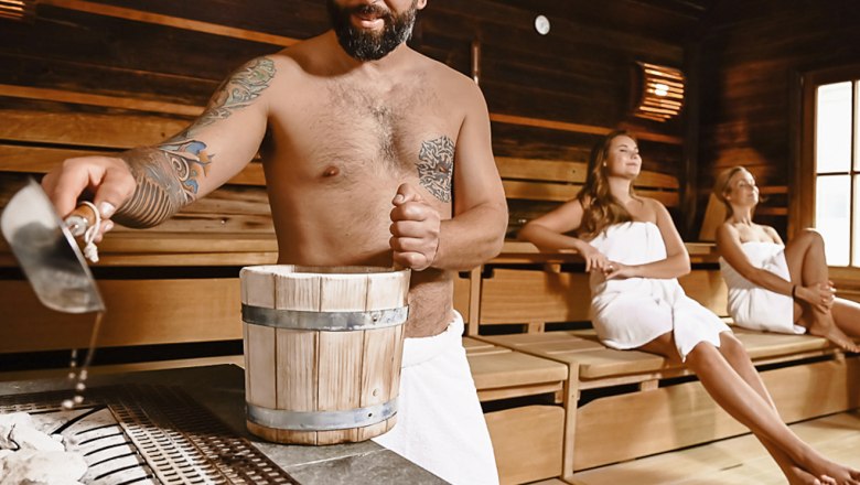 Mann gießt Wasser in Sauna, zwei Frauen sitzen entspannt.