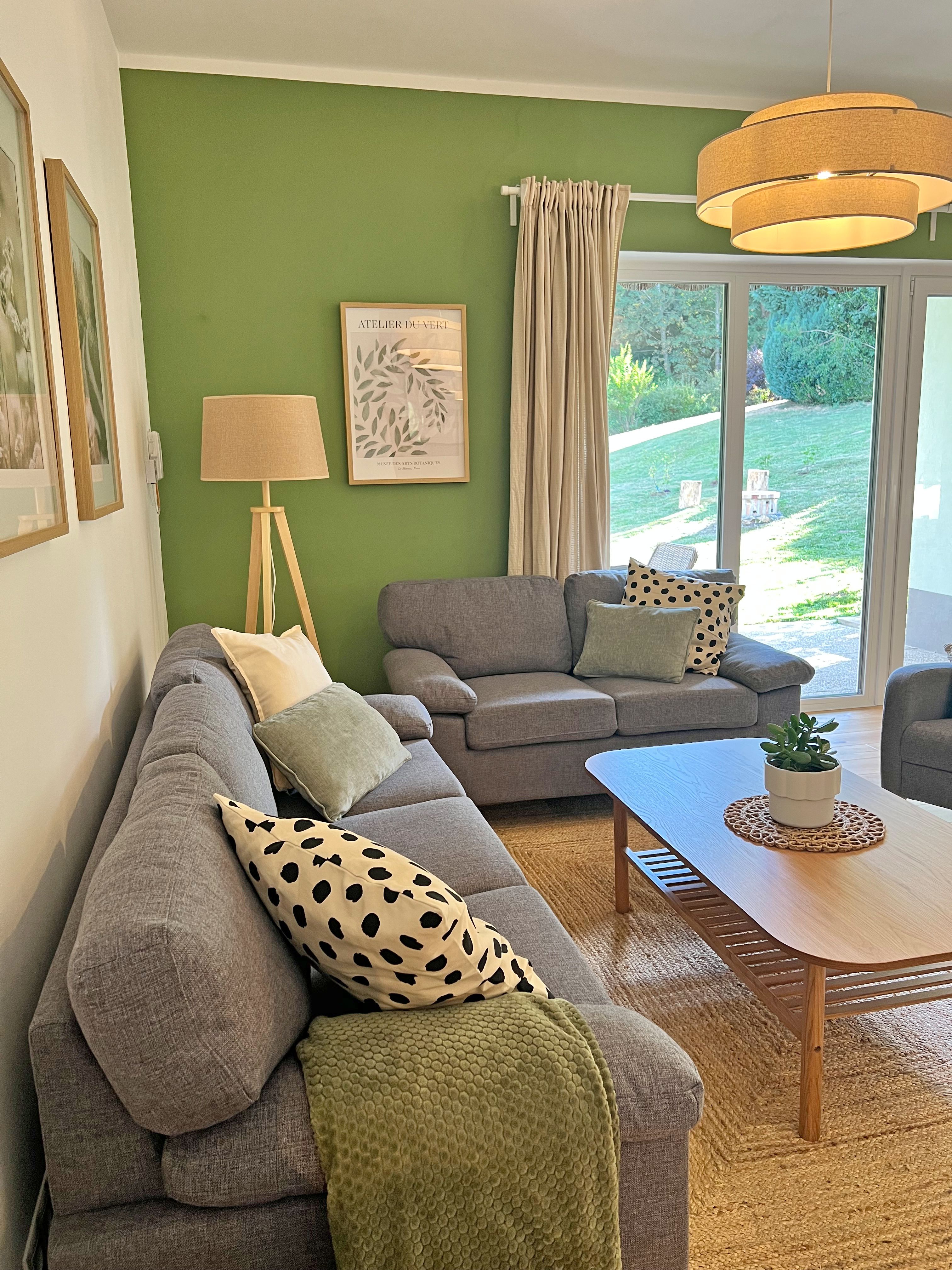 A cozy living room with a green accent wall, gray sofas, a wooden table, a floor lamp, and a view of the garden.