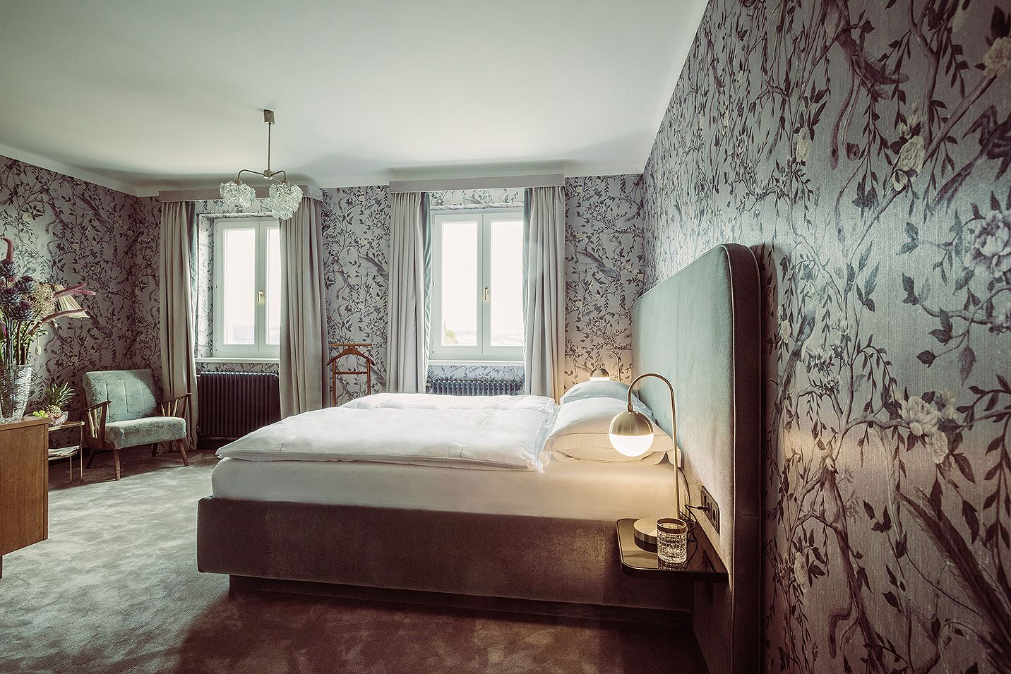 Elegantes Schlafzimmer mit floral gemusterten Tapeten, großem Bett und zwei Fenstern.