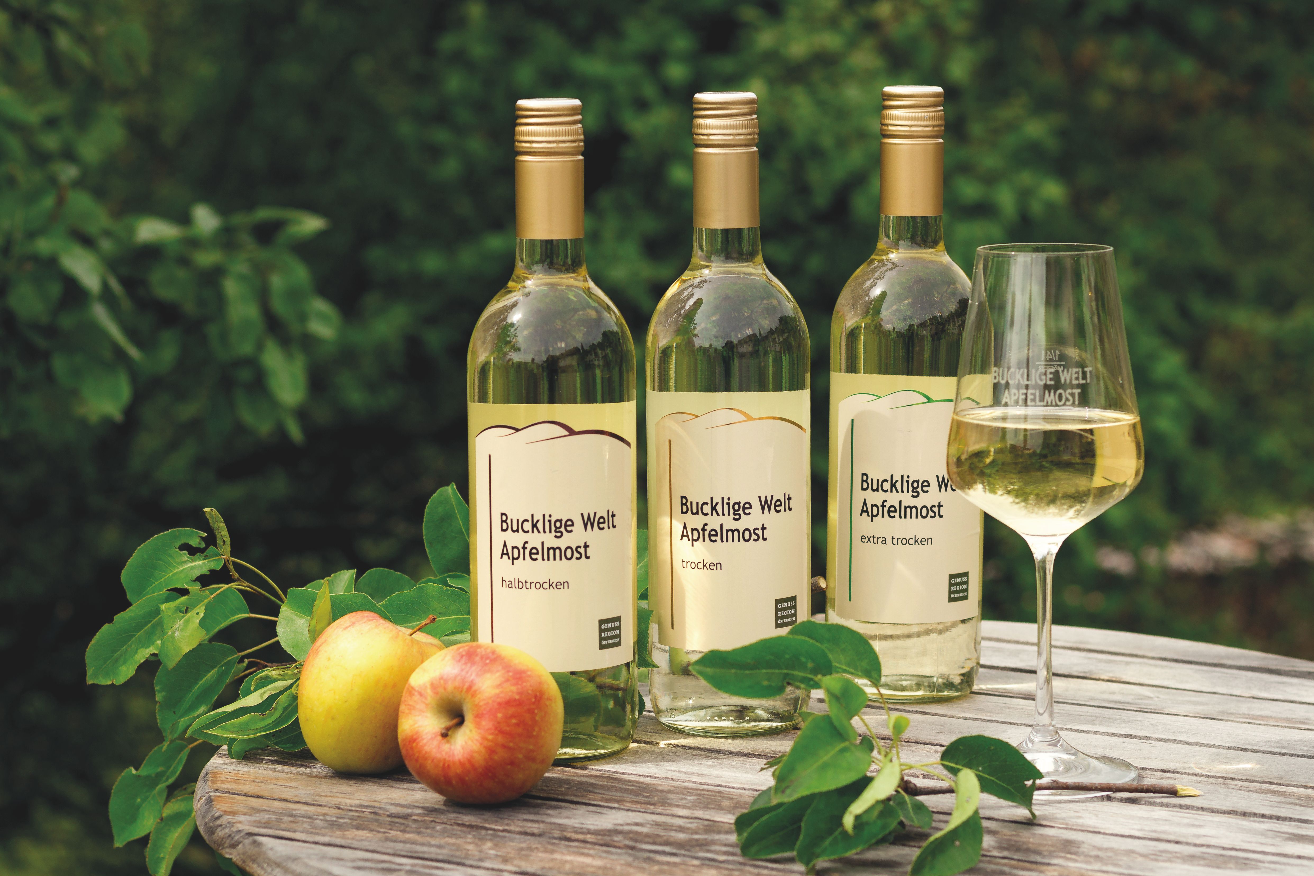 Drei Flaschen Apfelmost, ein Glas mit Apfelmost, Zweige und Äpfel auf einem runden Holztisch.