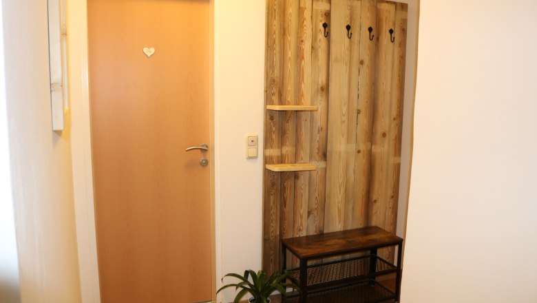Eingangsbereich mit Holztür, Garderobe aus Holz mit Haken, Bank und Pflanze.
