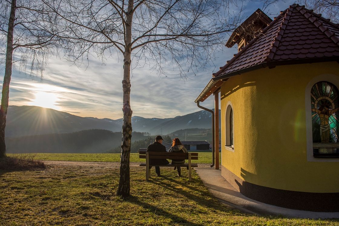 Zwei Personen sitzen auf einer Bank neben einer kleinen Kapelle mit Blick auf die Berge bei Sonnenuntergang.