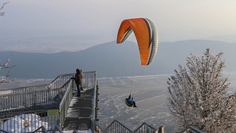 Ein Paraglider fliegt über eine verschneite Landschaft im Naturpark Hohe Wand, während eine Person auf einer Aussichtsplattform steht.
