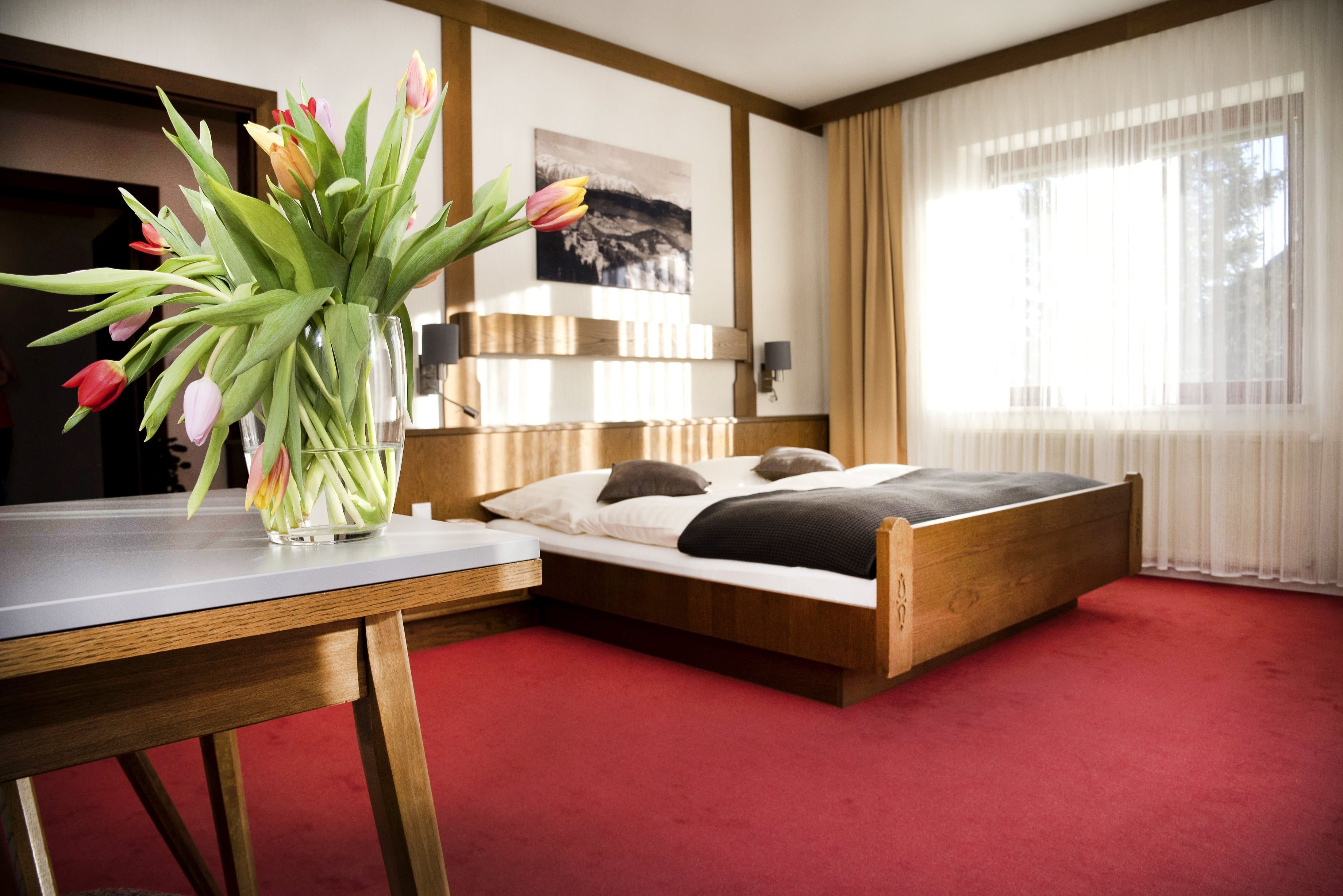 Ein gemütliches Doppelzimmer mit einem Holzbett, roten Teppichboden und einem Tisch mit einem Blumenstrauß aus Tulpen.