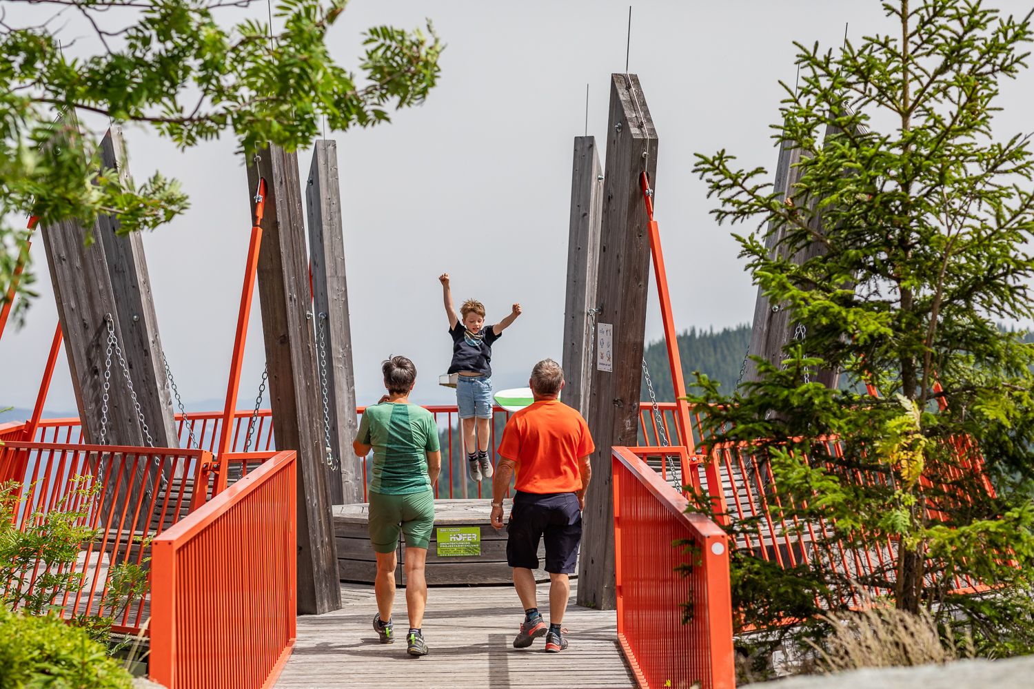 Ein Kind schaukelt auf einer Aussichtsplattform mit roten Geländern, während zwei Erwachsene zuschauen.