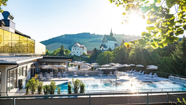 Außengelände eines Hotels mit Pool, Sonnenschirmen und Liegen, umgeben von Natur und Kirche im Hintergrund.