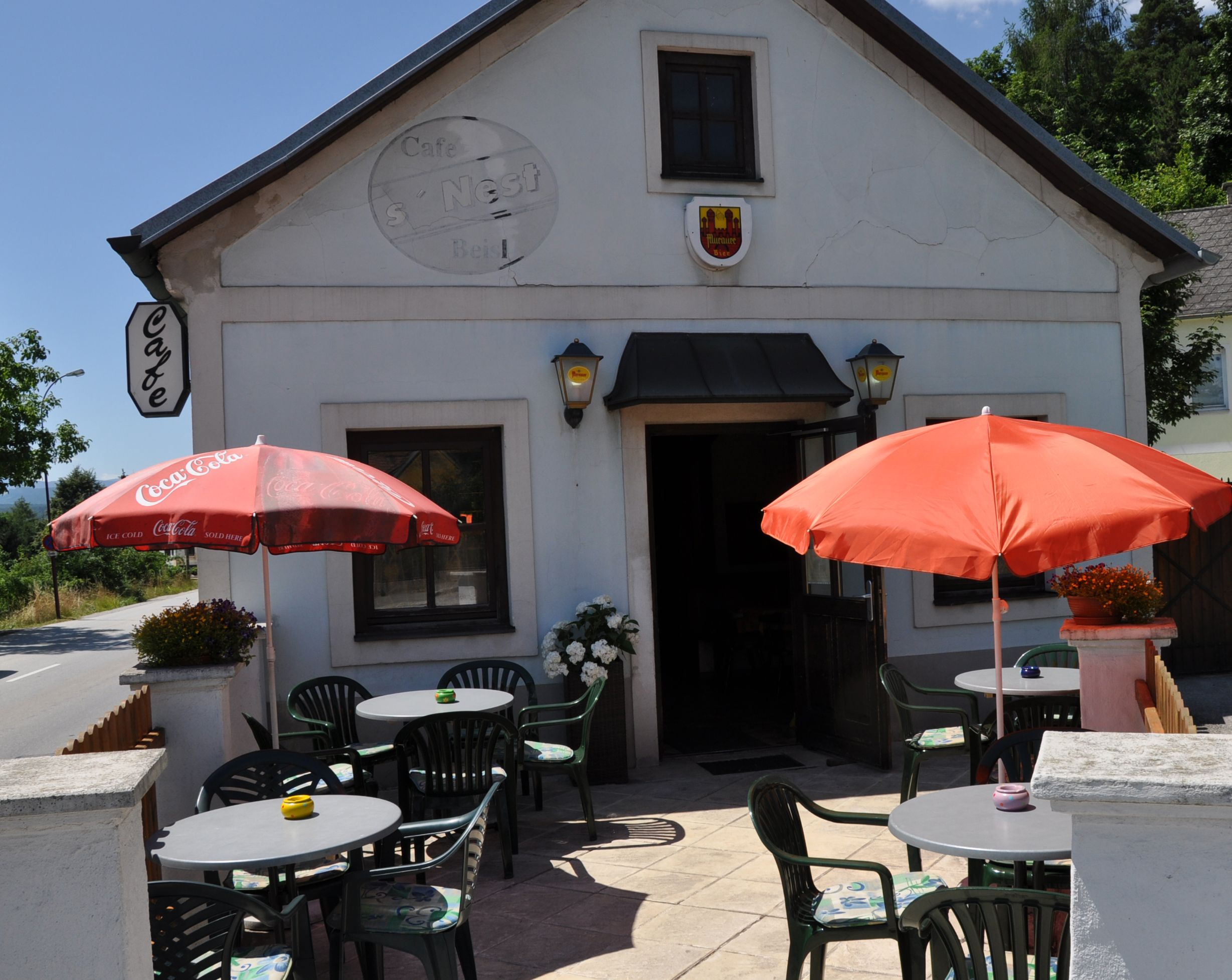 Außenansicht eines Cafés mit Terrasse, Tischen und Sonnenschirmen.