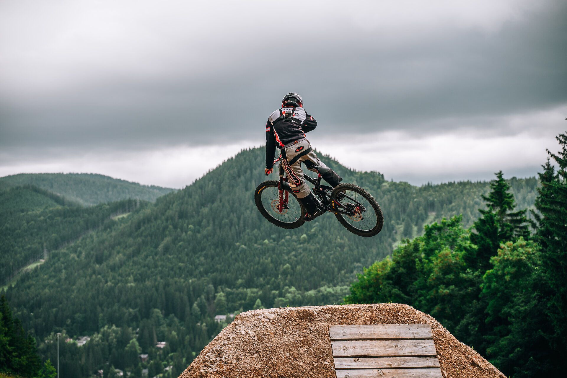 Mountainbiker springt mit dem Fahrrad von einem Holzabsprung vor einer grünen Berglandschaft.
