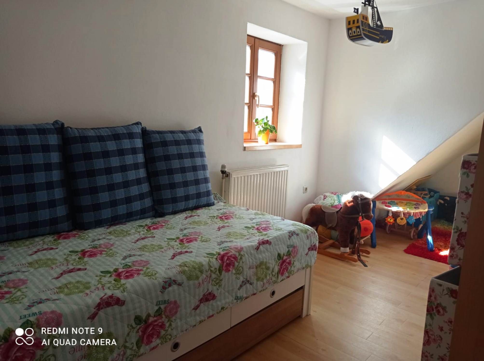 Kinderzimmer mit ausziehbarem Bett, karierten Kissen, Spielzeugpferd und Spieltisch. Helles Fenster mit Pflanze.