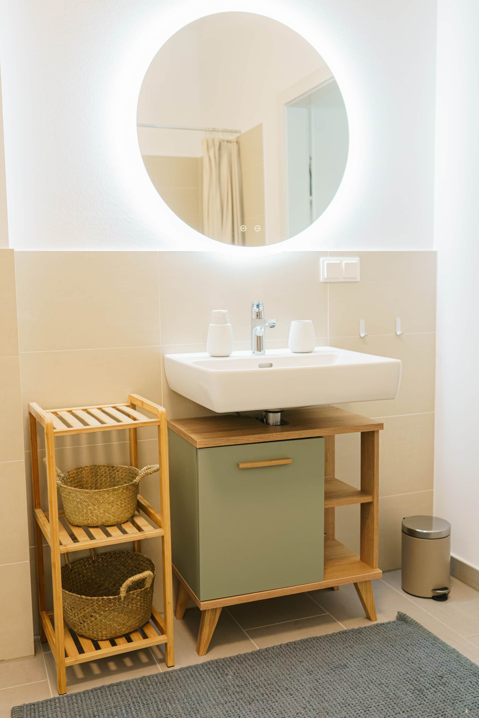 Badezimmer mit Waschbecken, salbeigrünem Unterschrank, Regal, Spiegel, Abfalleimer