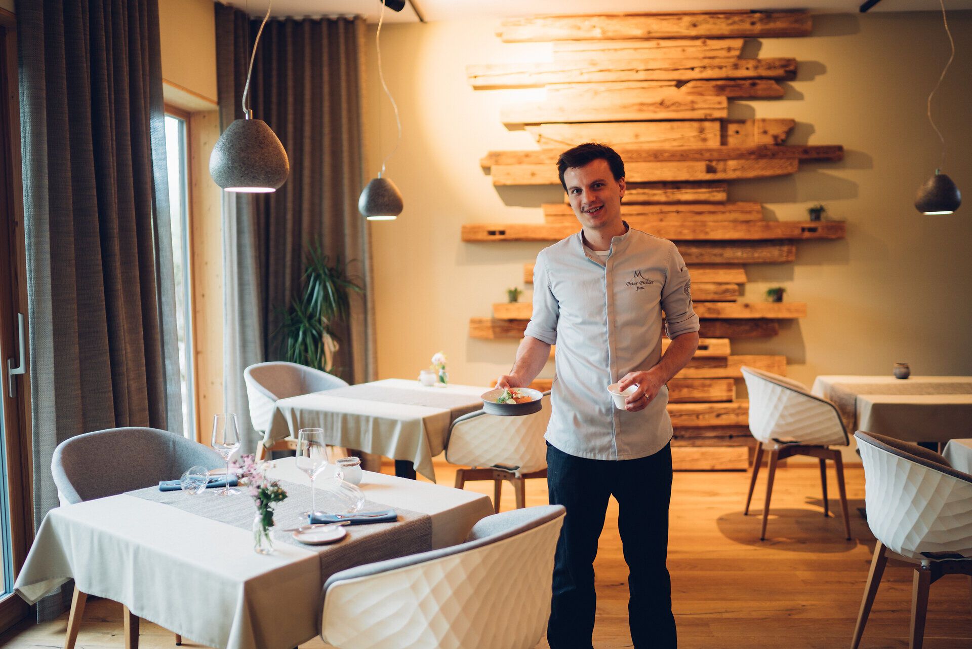 In einem stilvollen Restaurant, umgeben von warmem Holz und sanften Farben, empfängt ein freundlicher Koch die Gäste mit einem kunstvoll angerichteten Gericht. Die Atmosphäre ist einladend und verspricht ein unvergessliches kulinarisches Erlebnis inmitten der malerischen Wiener Alpen.