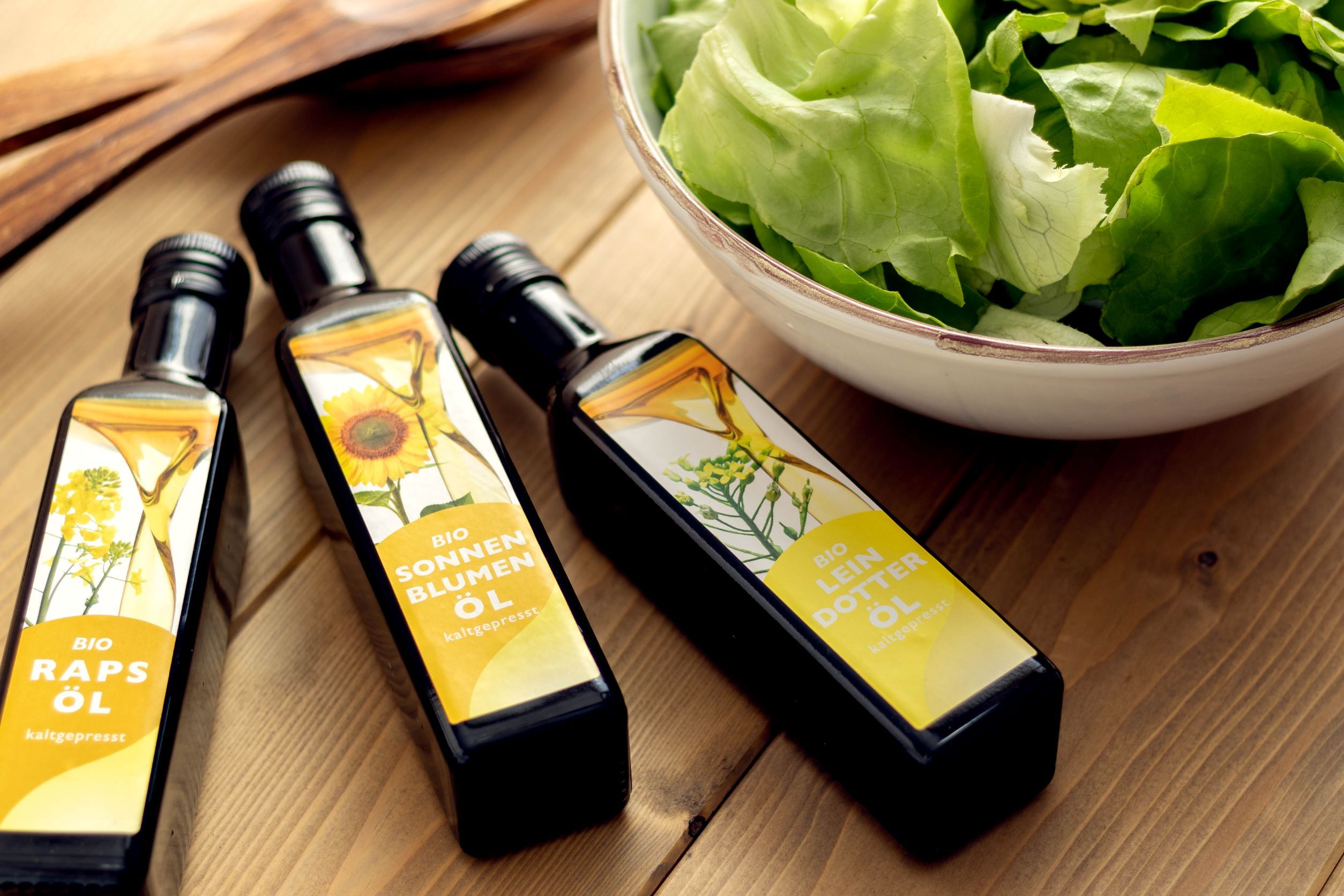 Drei verschiedene Öle liegen auf einem Holztisch neben einer Schüssel mit grünem Salat.