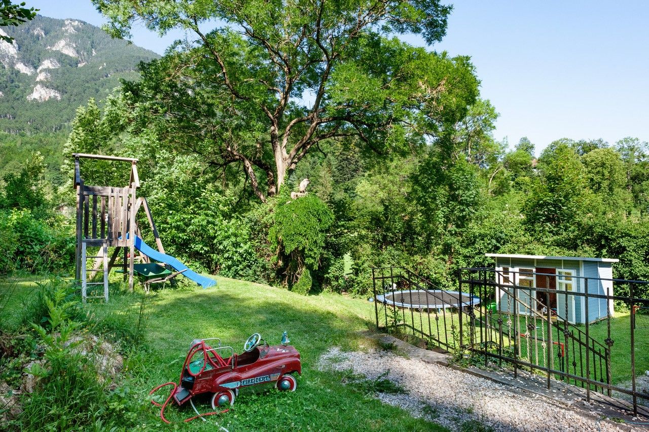 Ein Garten mit Spielplatz, Rutsche, Spielzeugauto und Trampolin vor einem Hintergrund aus Bäumen und Bergen.