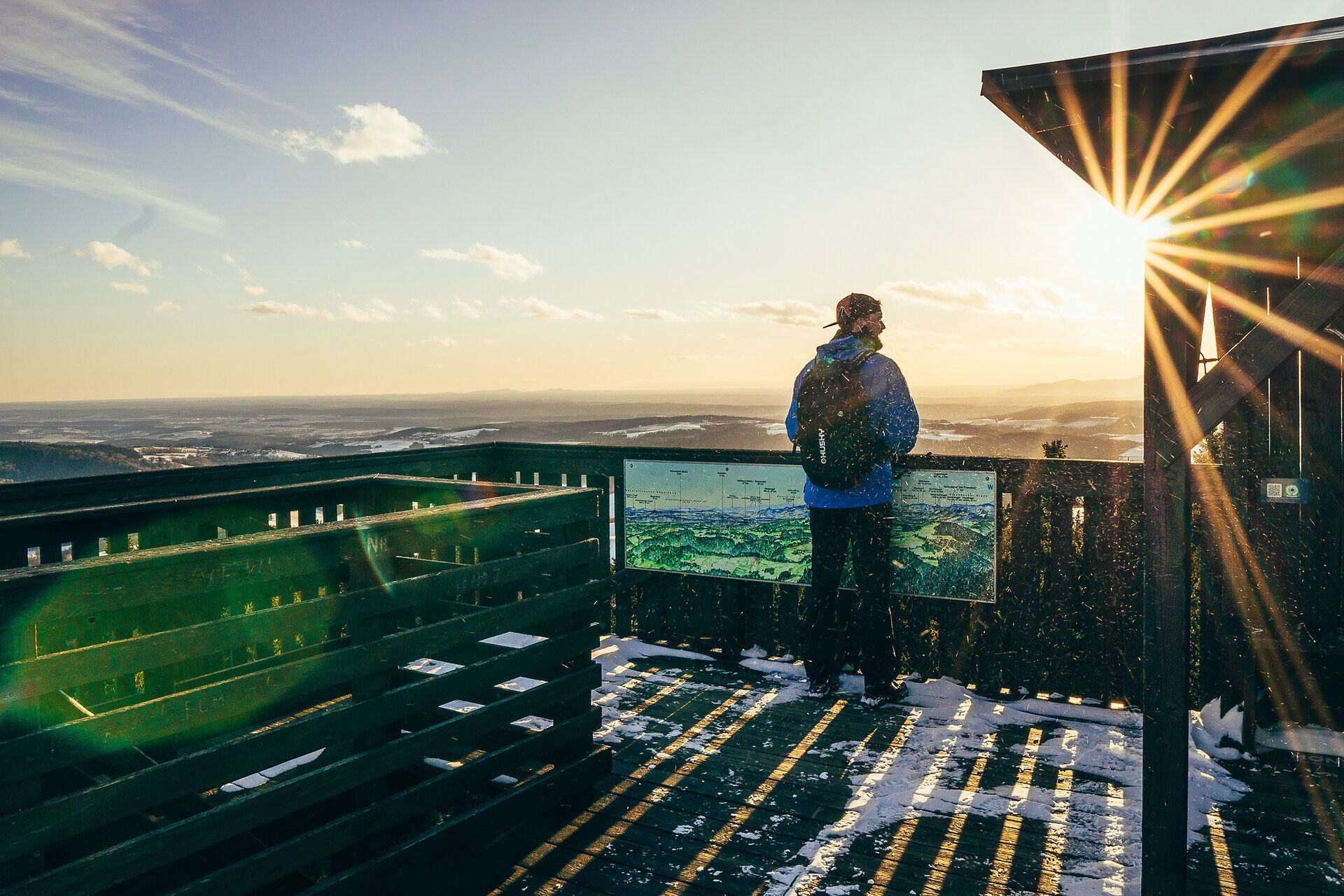 Die winterliche Landschaft erstreckt sich weit und breit, während die Sonne sanft über den schneebedeckten Hügeln aufgeht. Ein Besucher genießt den atemberaubenden Ausblick und lässt sich von der friedlichen Atmosphäre der Buckligen Welt verzaubern.