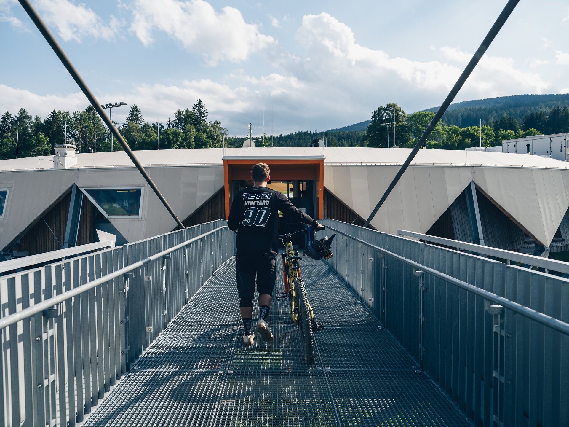 Ein Mountainbiker genießt die frische Bergluft, während er auf einer schmalen Brücke in Richtung des Bikeparks fährt. Umgeben von üppigem Grün und majestätischen Bergen, spürt er die Freiheit und das Abenteuer, das die Wiener Alpen in Niederösterreich bieten.