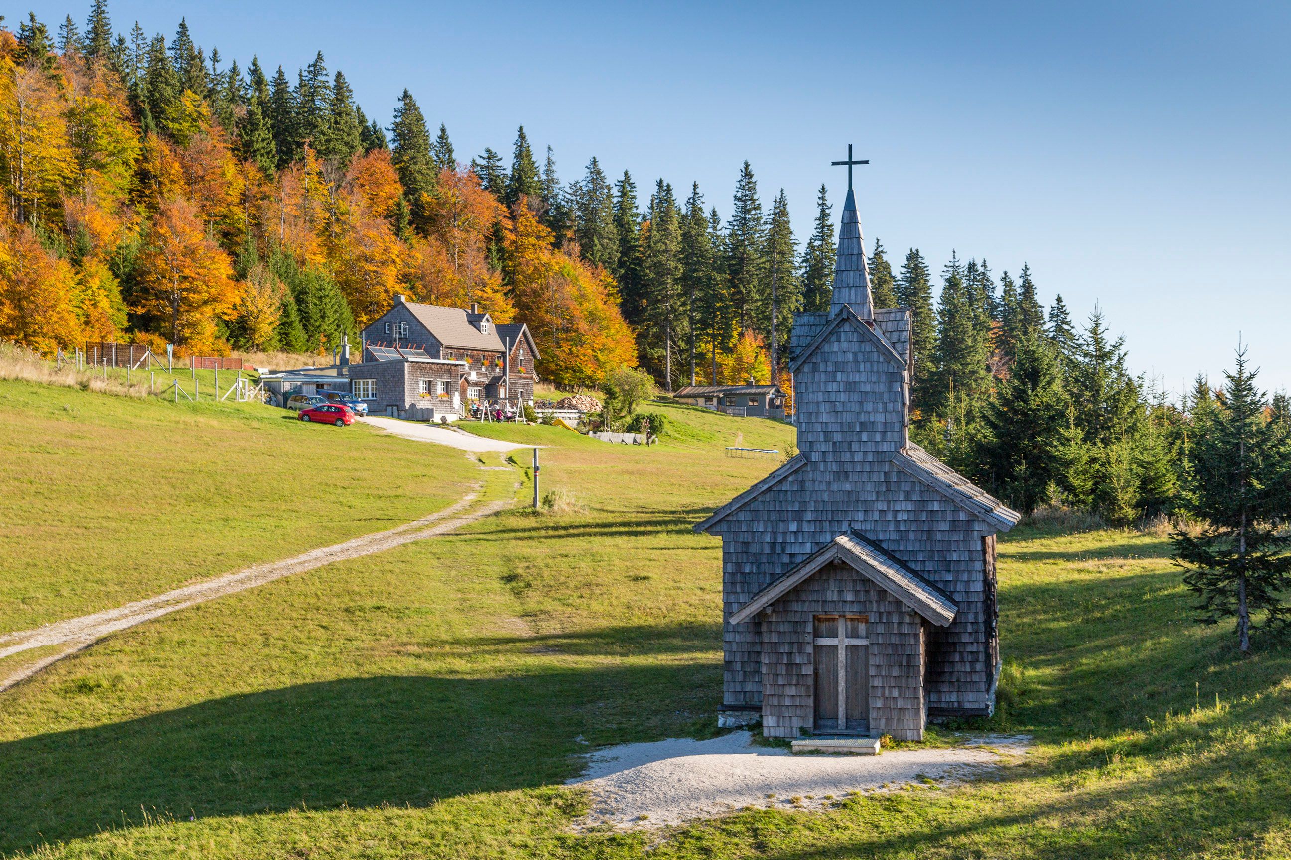 A wooden church at the summit of the Unterberg with a cross, with meadows and a hiking trail in front of it; in the background, an autumn forest and a mountain lodge.