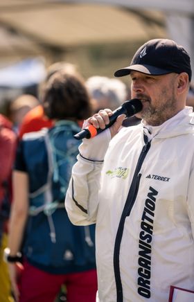 Ein Mann in einer weißen Sportjacke und schwarzer Kappe spricht in das Mikrofon, dass er in der Hand hält. Auf der Jacke steht in schwarz Organisation.