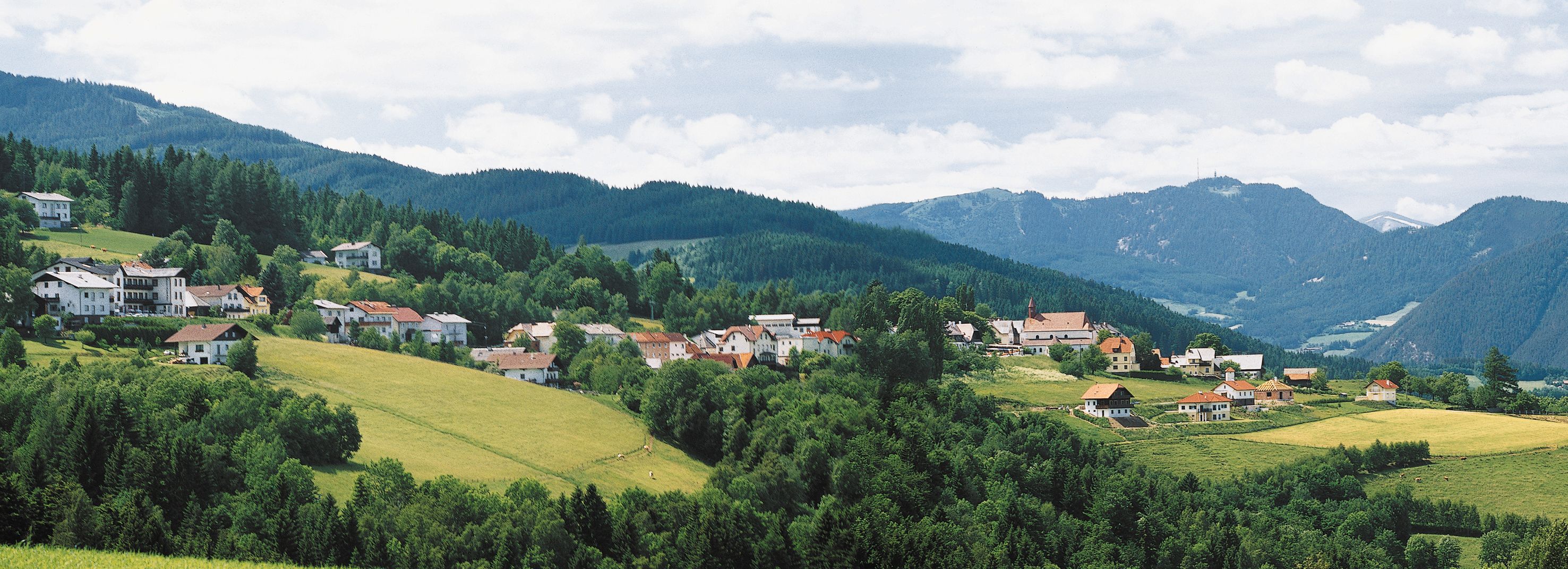 Panorama von St. Corona am Wechsel mit grünen Hügeln und Bergen im Hintergrund.