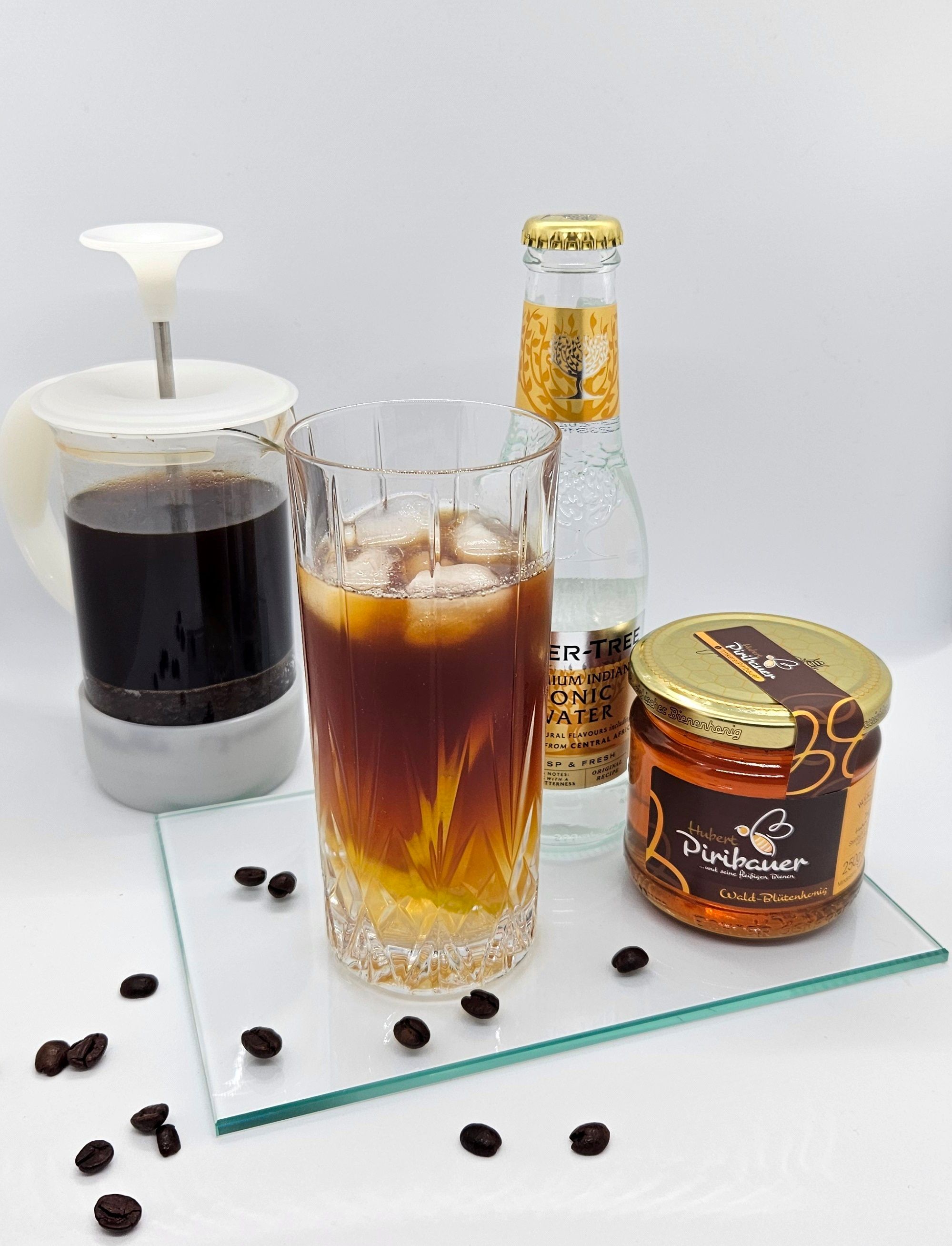 Ein Glas Cold-Brew-Tonic mit Eiswürfeln, daneben Honigglas, Glasflasche und Kaffeekanne auf Glasplatte.