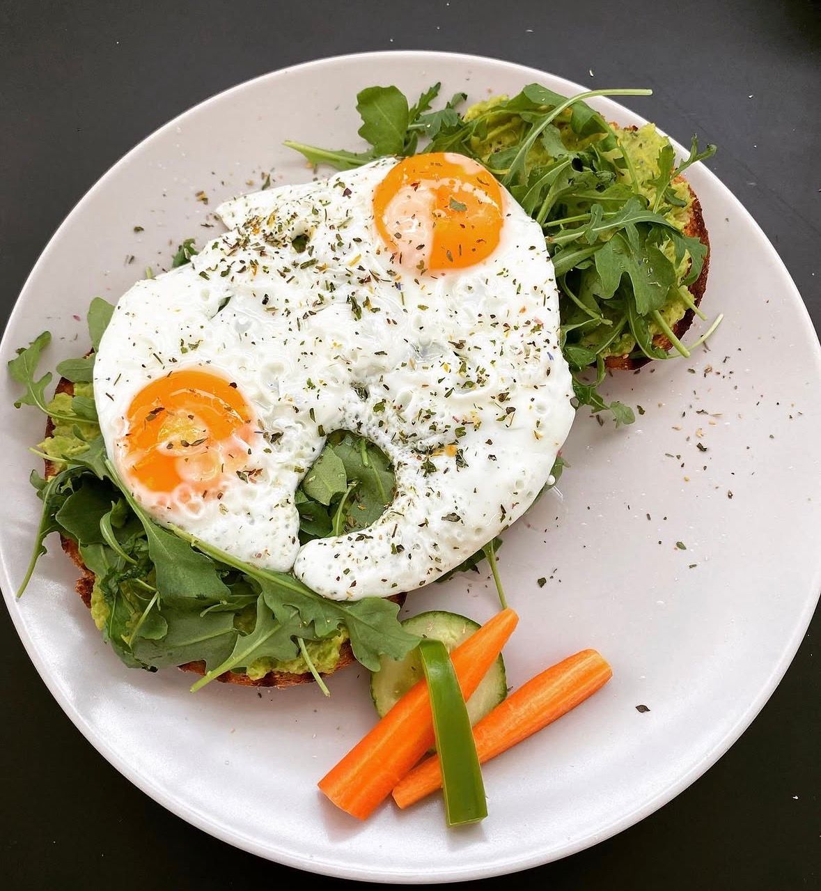 Zwei belegte Brote mit Avocado, Rucola und Spiegeleiern auf einem weißen Teller, dazu etwas Gemüse.