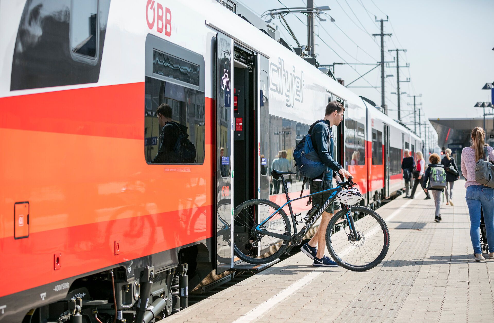 Die Sonne strahlt über den Bahnhof, während Reisende mit ihren Fahrrädern in den Zug einsteigen. Die frische Luft und die Vorfreude auf neue Abenteuer in der Natur sind spürbar. Ein perfekter Start für eine unvergessliche Reise in die Berge.