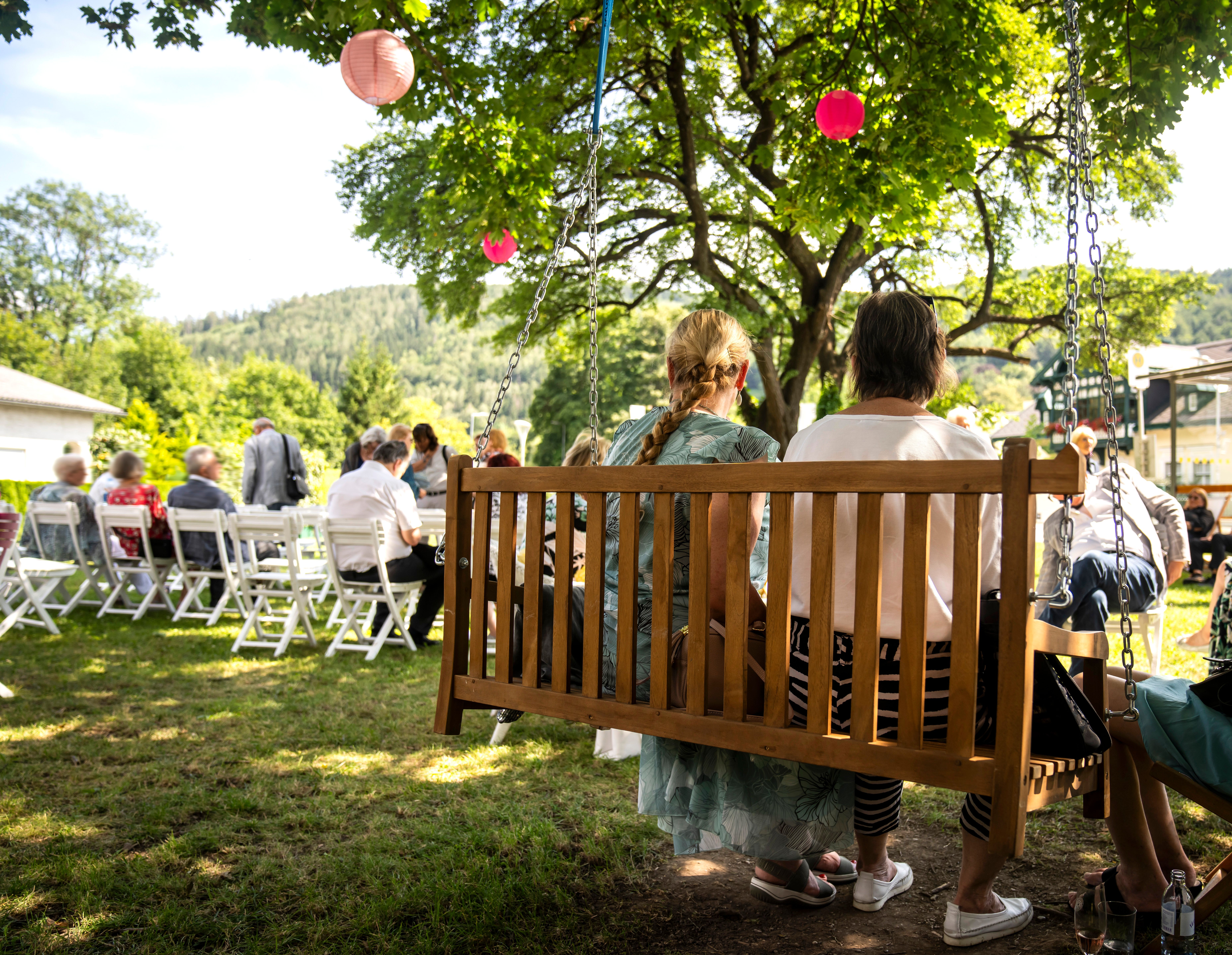Unter dem schattenspendenden Baum genießen Besucher eine entspannte Zeit auf einer Holzbank, während im Hintergrund fröhliche Gespräche und Lachen von den Festspielen erklingen. Die sanfte Brise und die bunten Laternen verleihen der Atmosphäre einen festlichen Charme, der die Schönheit des Sommers in den Wiener Alpen widerspiegelt.