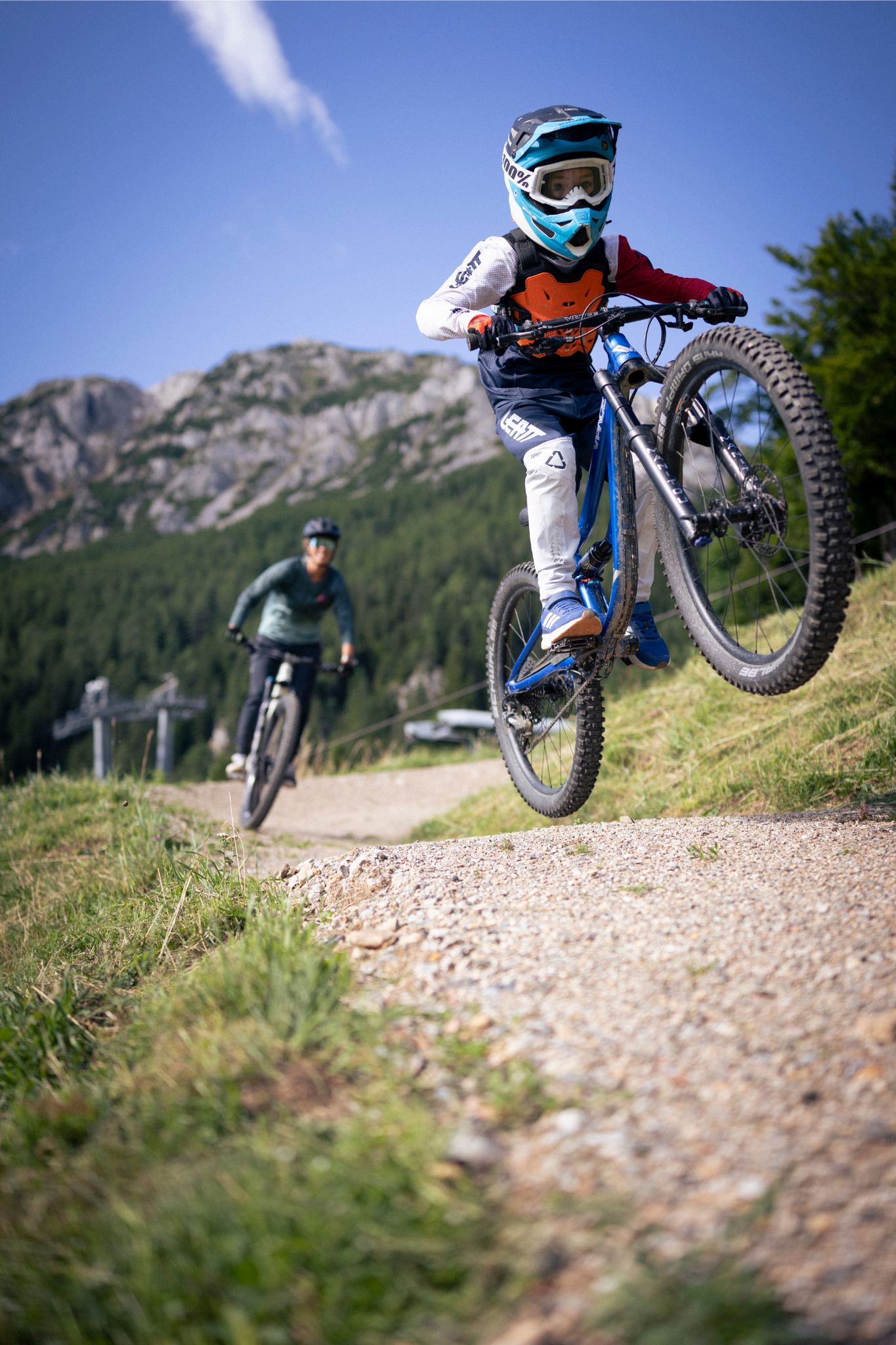 Ein Kind auf einem Mountainbike springt auf einem Schotterweg, gefolgt von einem Erwachsenen. Im Hintergrund sind Berge und Wälder zu sehen.