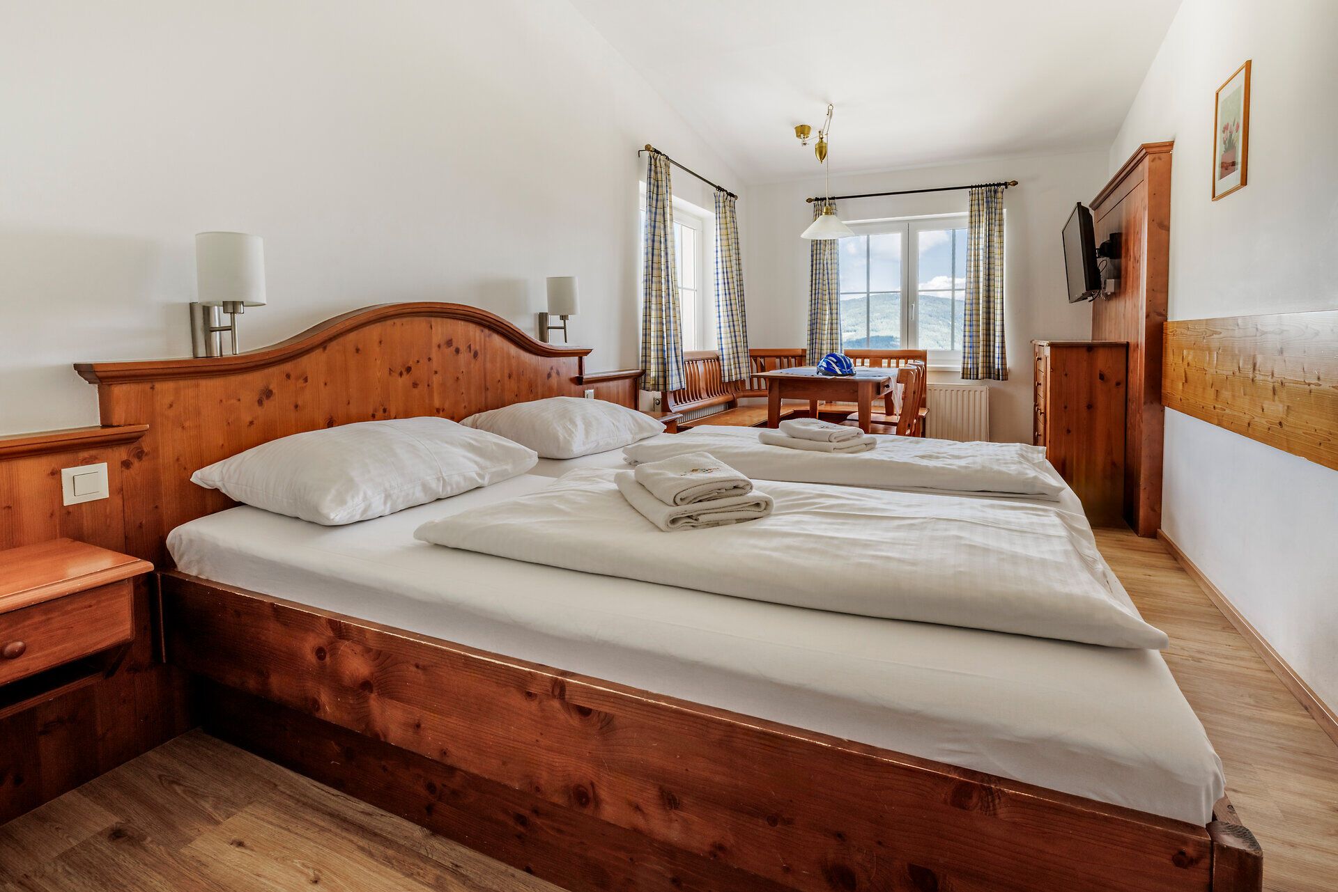 Der Blick in das Zimmer im Alpengasthof Enzian zeigt einen Teil der Ausstattung: Doppelbett mit weißer Bettwäsche und Handtüchern, Sitzecke vor zwei Fenstern, Fernseher über einer Kommode.ausgestatten mit einem Doppelbett, einer Sitzecke sowie einem Fernseher über einer Kommode.