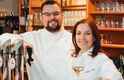Ein Mann und eine Frau in weißen Kochjacken stehen hinter einer Schank aus Holz.