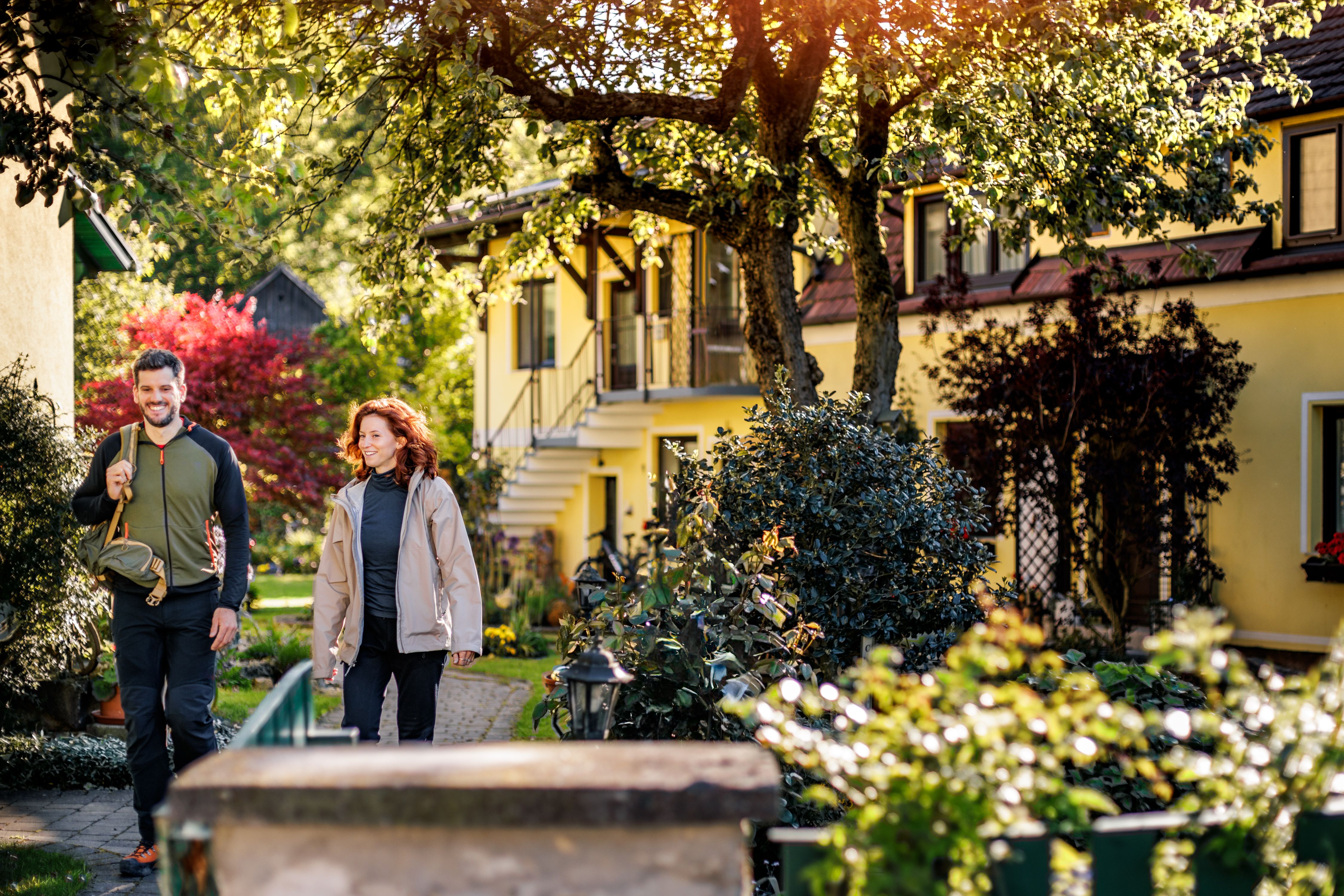 Ein Mann und eine Frau spazieren lächelnd durch einen sonnigen Garten mit gelben Häusern im Hintergrund.