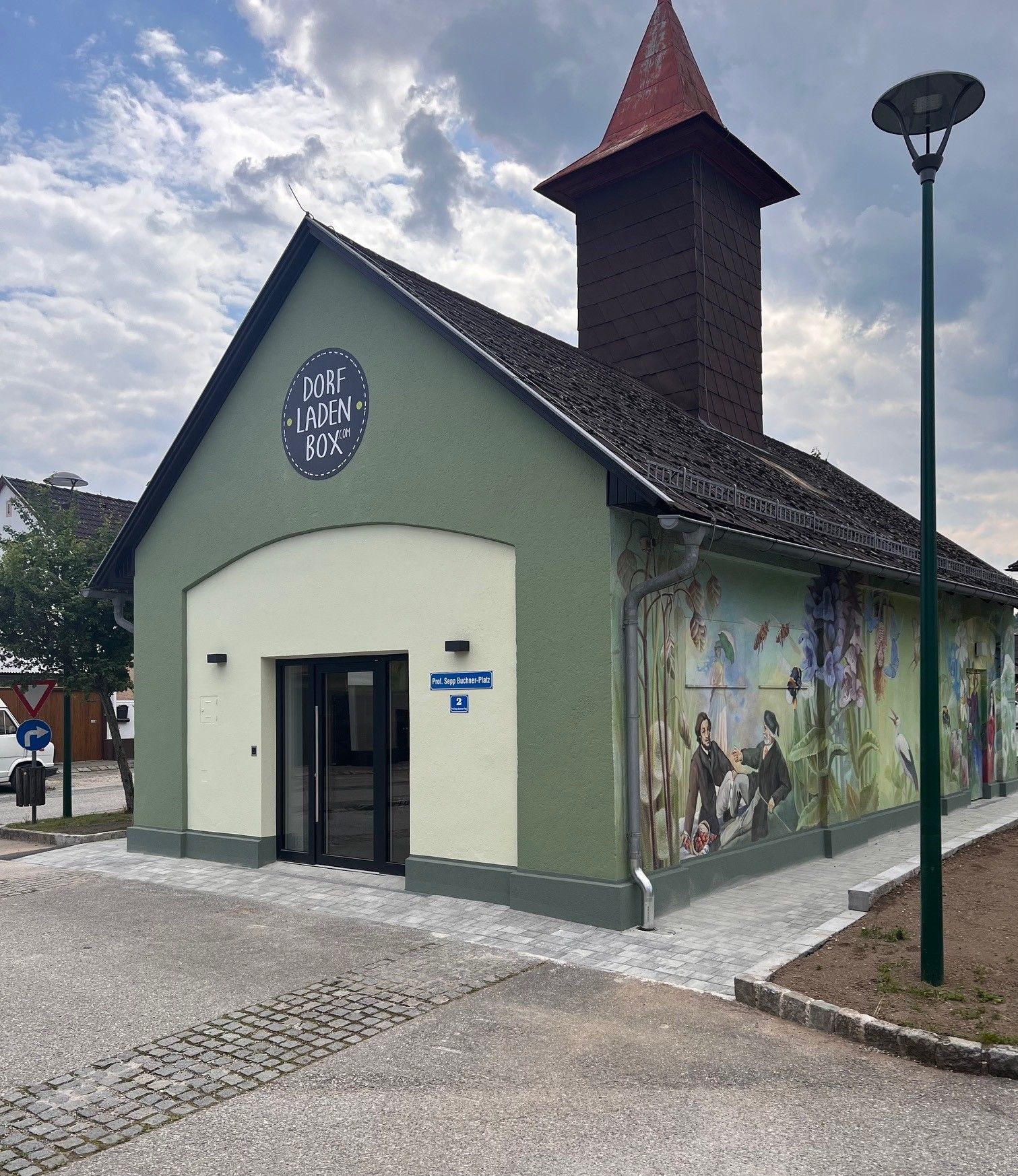 Ein kleines, grünes Gebäude mit der Aufschrift 'Dorfladen Box' und einem gemalten, ländlichen Wandbild.