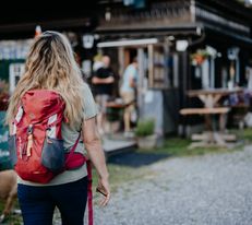 Eine Wanderin mit rotem Rucksack geht auf die Holzhütte zu.