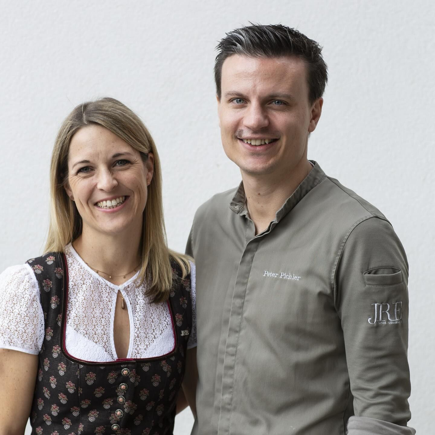 Ein Mann in Kochjacke und eine Frau im Dirndl vor einer weißen Wand. 