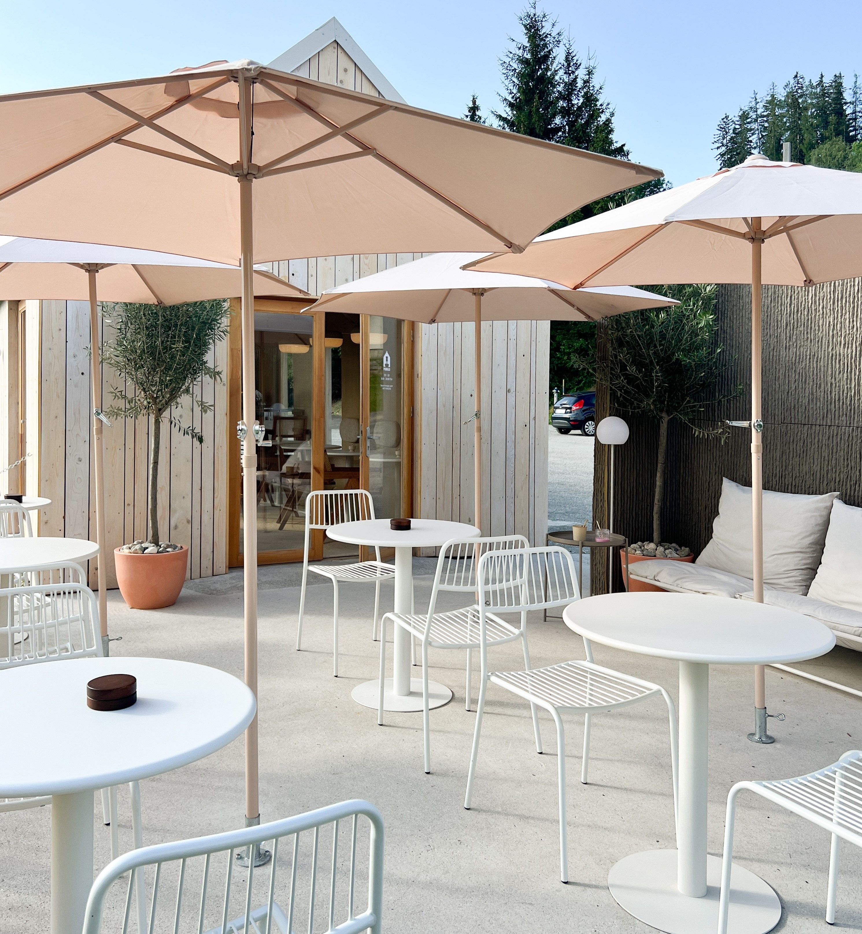 Moderne Terrasse mit weißen Tischen und Stühlen, Sonnenschirmen und Holzhütte im Hintergrund.