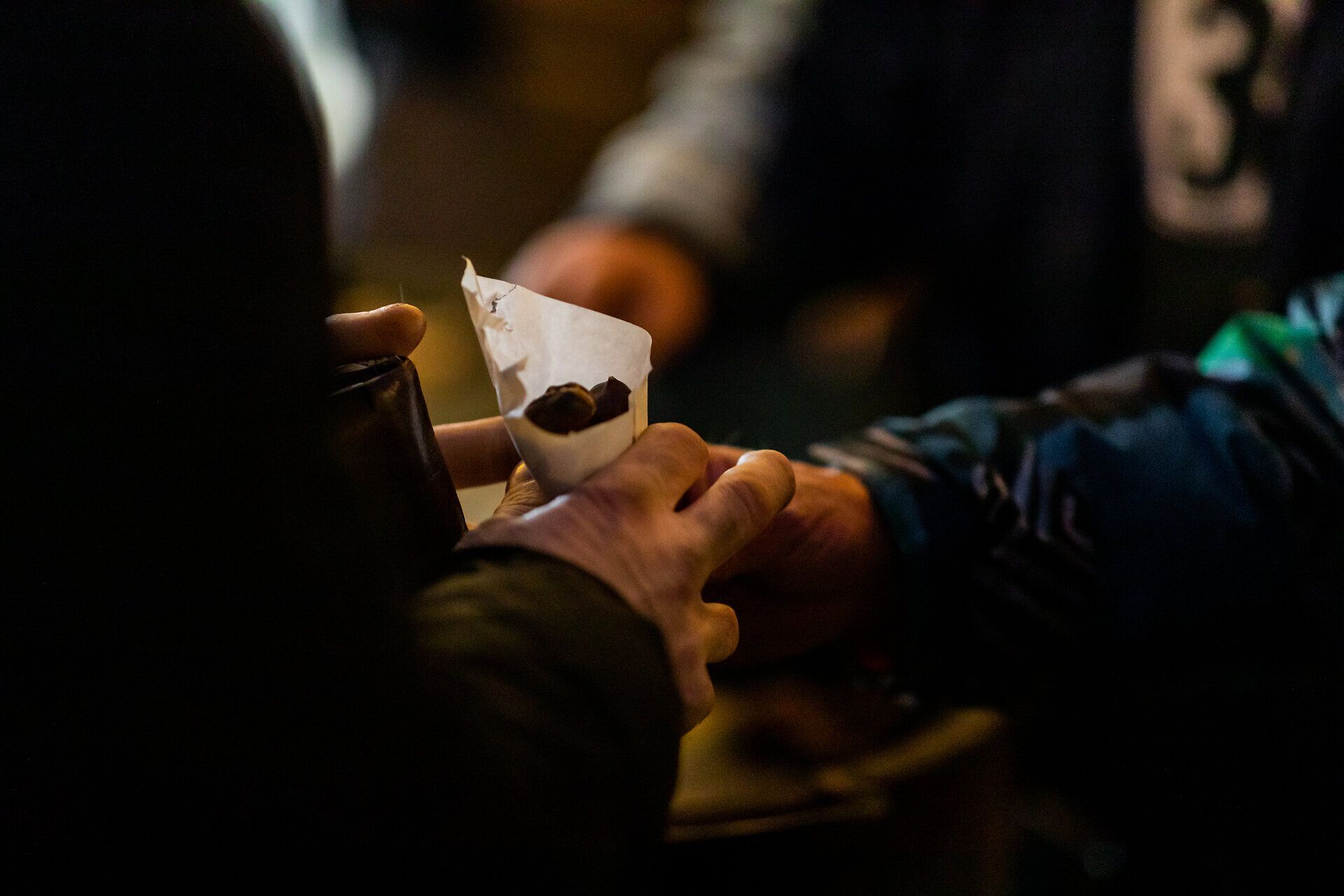 Hände halten eine Tüte mit Maroni auf einem Weihnachtsmarkt bei Nacht.
