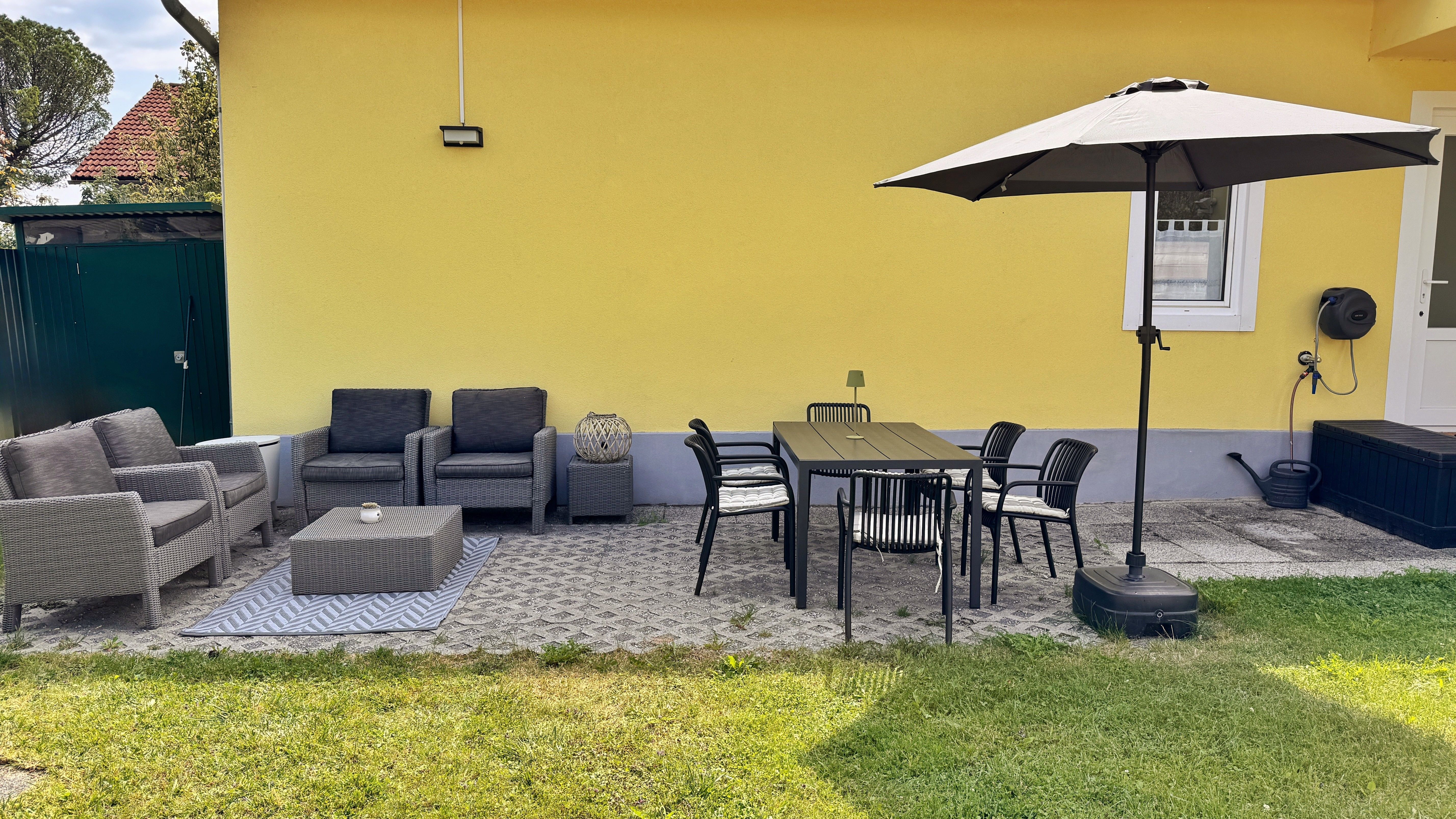 Terrasse mit gelber Hauswand, grauen Sitzmöbeln, Esstisch, Stühlen und Sonnenschirm.