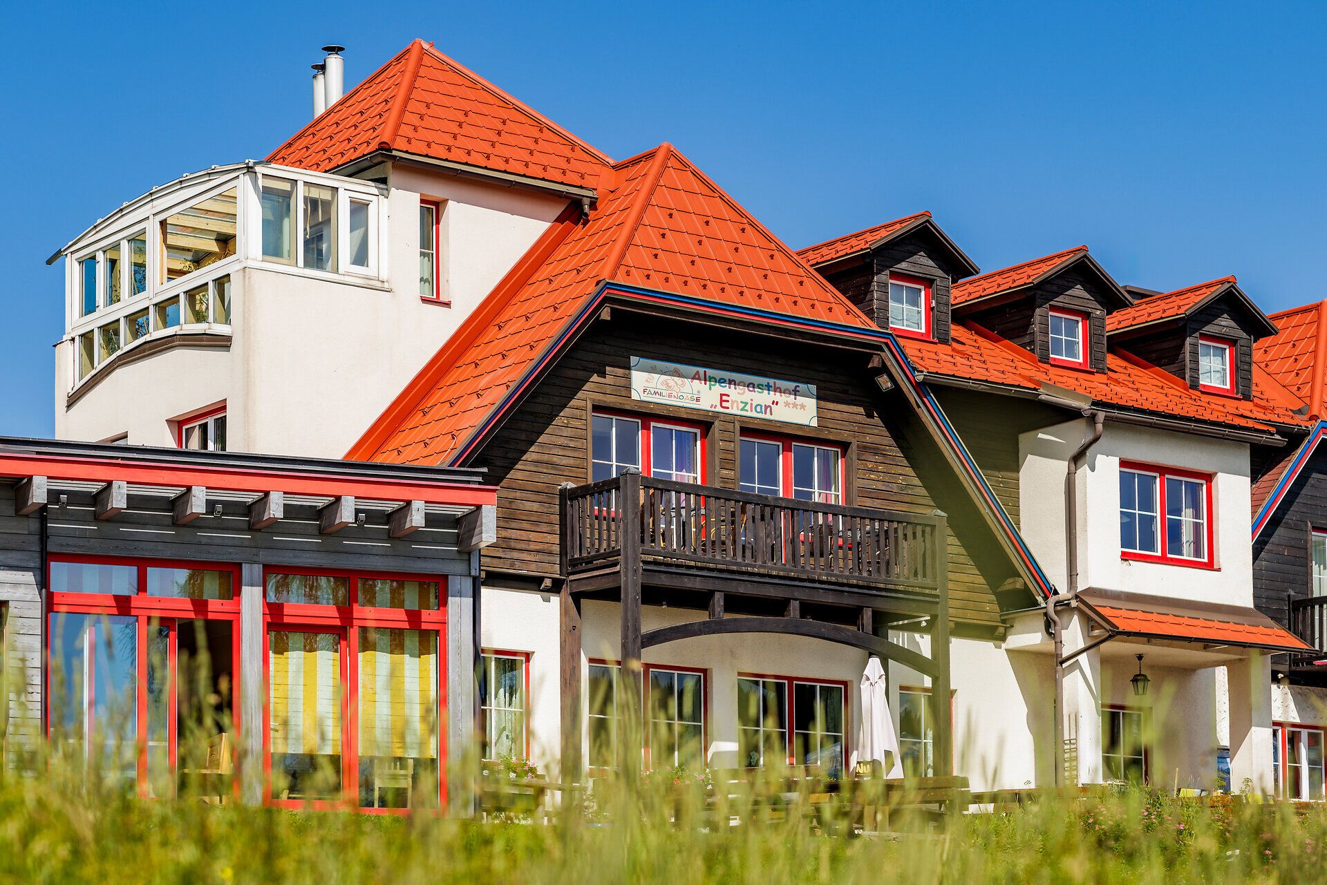 Nahansicht vom Alpengasthof Enzian mit heller Fassade, Holzfassade und rot-weißen Fenstern. Hinter dem Holzbalkon ist die Fassade holzvertäfelt und trägt die bunte Aufschrift "Alpengasthof Enzian" auf weißem Hintergrund.