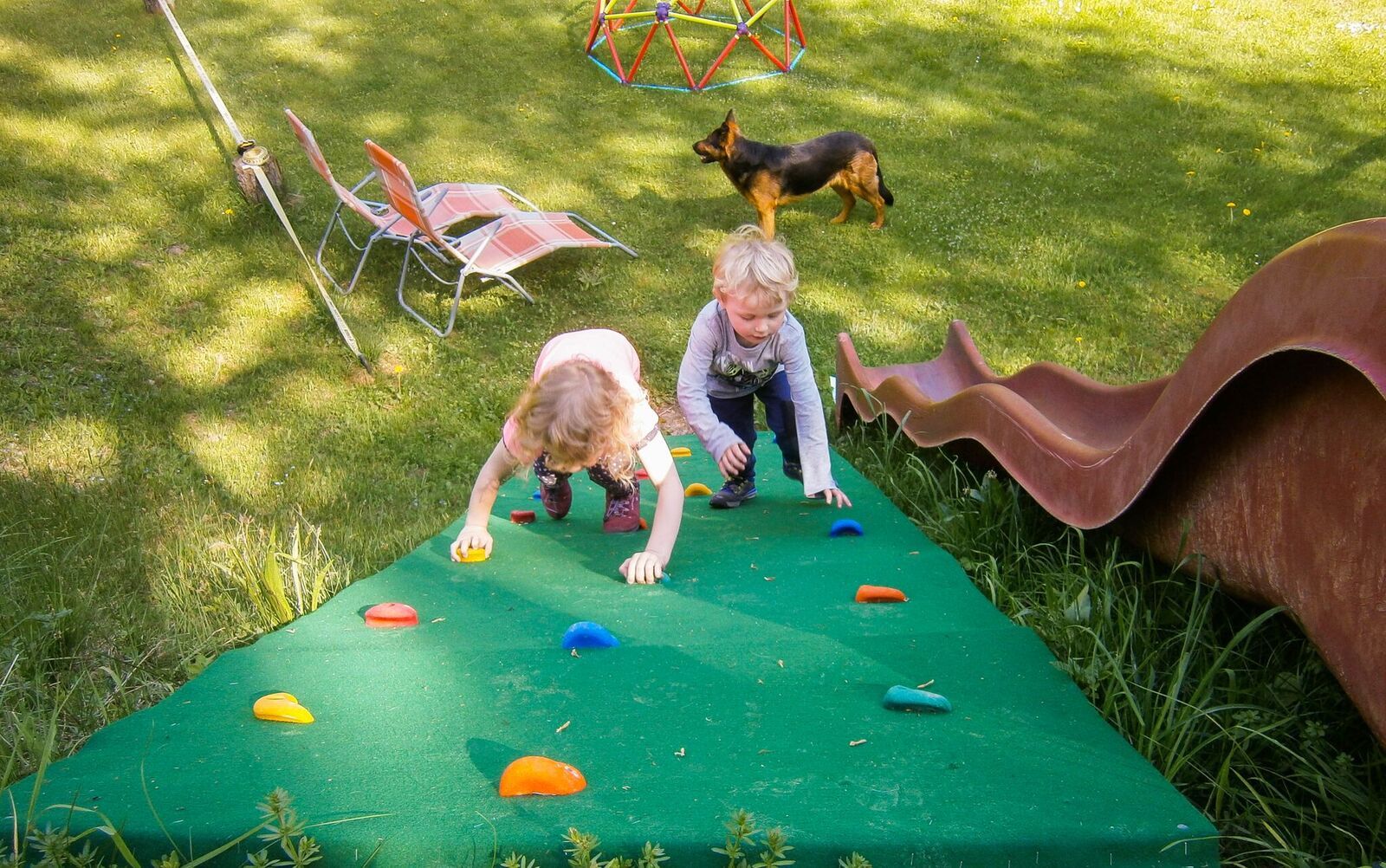 Zwei Kinder klettern auf einem Spielplatz eine grüne Kletterwand hinauf, während ein Hund im Hintergrund steht.