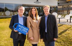 Drei Personen stehen vor dem Kulturhaus, einem modernen Gebäude mit Glasfront und der Aufschrift "Sconarium Bad Schönau". Der Bürgermeister hält ein das Logoschild "Wiener Alpen" in den Händen. 