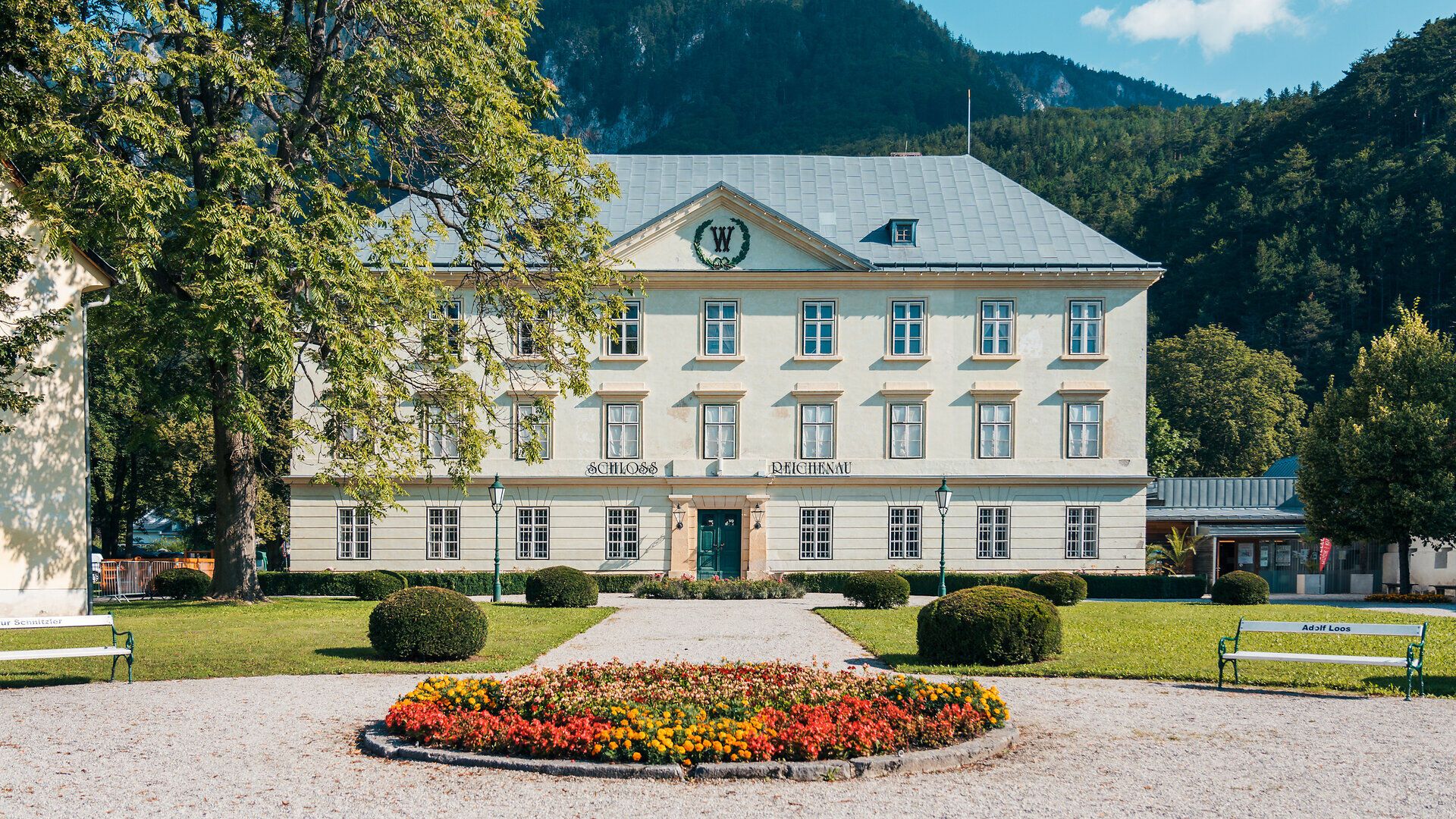 Historisches Schloss Reichenau mit symmetrischer Fassade, gepflegtem Garten und Blumenbeet vor bewaldeter Bergkulisse an einem sonnigen Tag.