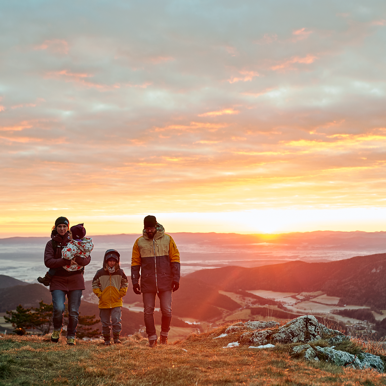 Die Familie genießt einen unvergesslichen Moment in der atemberaubenden Berglandschaft, während die Sonne hinter den sanften Hügeln untergeht. Die warmen Farben des Sonnenuntergangs tauchen die Umgebung in ein magisches Licht und laden zu weiteren Abenteuern in der Natur ein.