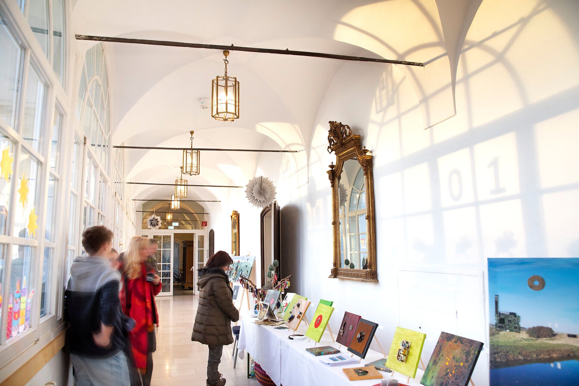 Heller Ausstellungsflur mit großen Fenstern, Besucher betrachten Kunstwerke auf Tischen.